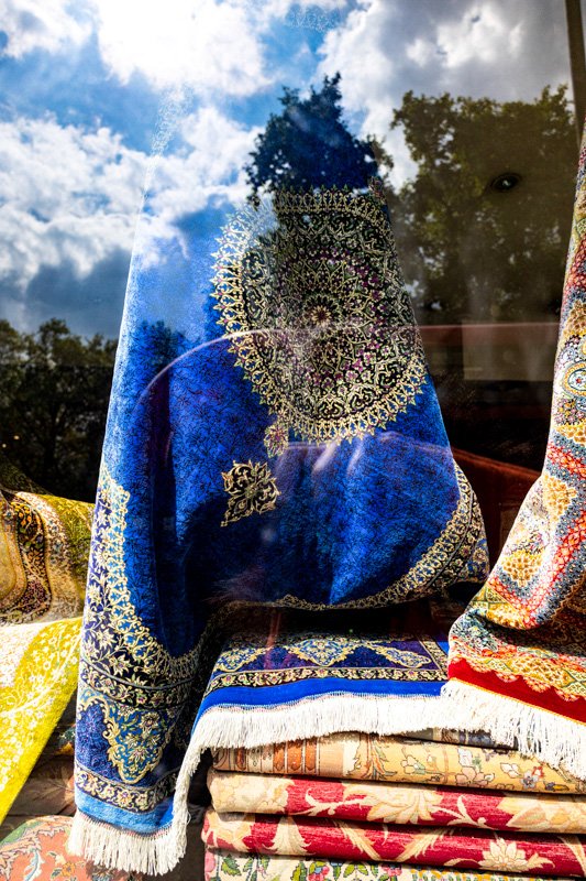 Display of colorful, intricately patterned Persian-style rugs and textiles behind a glass window, reflecting trees and a partly cloudy sky.