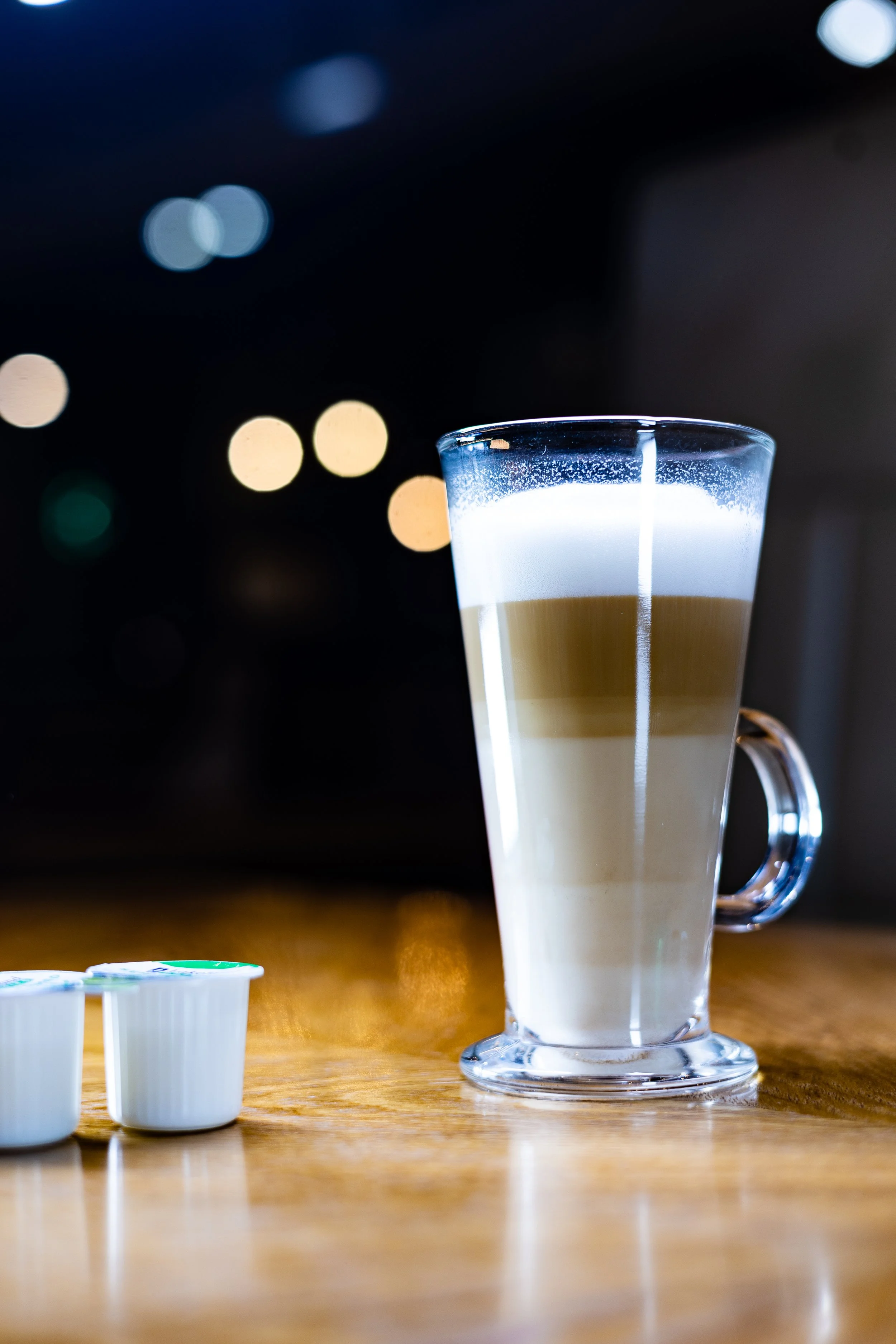 A layered latte drink in a clear glass mug on a wooden surface, with coffee creamer pods nearby and out-of-focus colorful lights in the background.