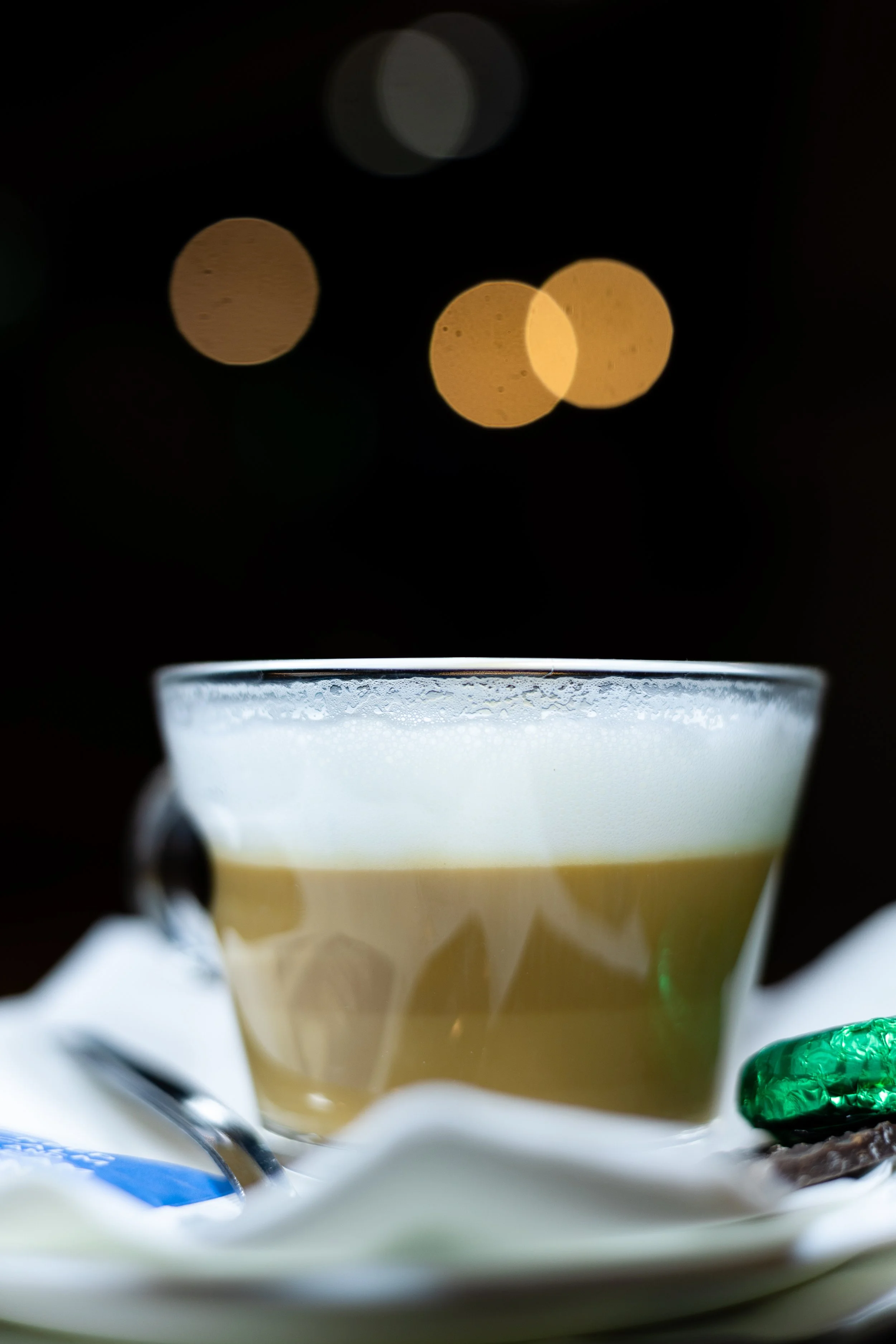 A glass cup filled with a layered, frothy coffee drink with a dark background and blurred lights in the distance.