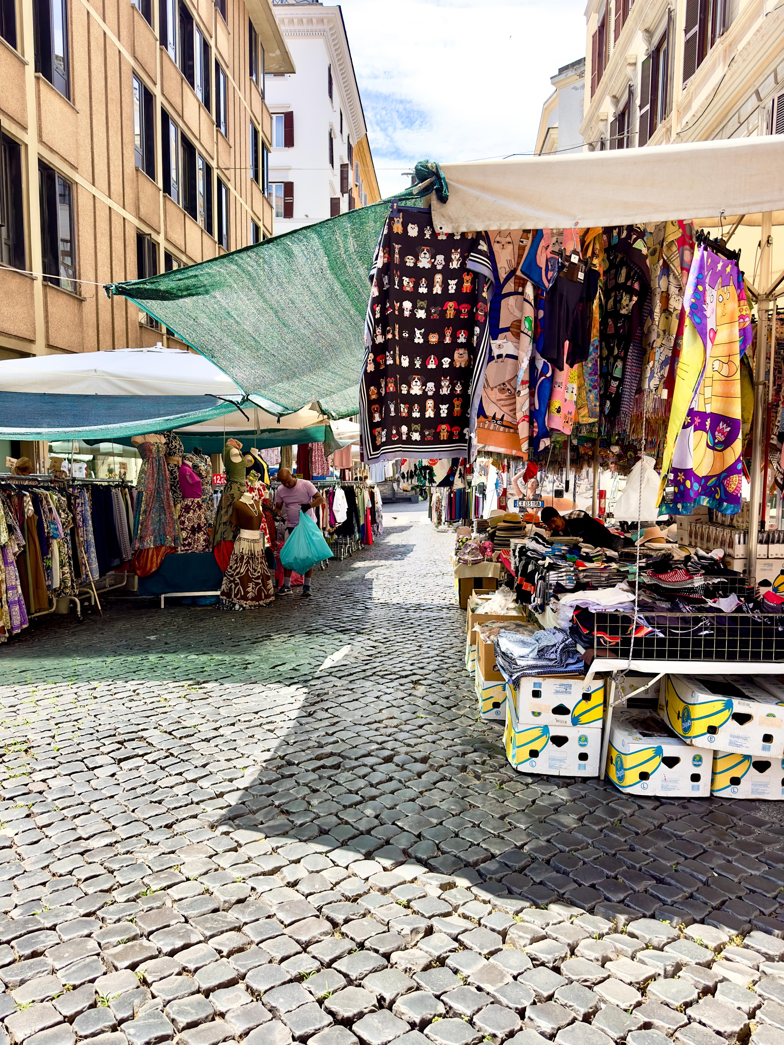 Outdoor street market with clothing and accessories on display, with cobblestone street and buildings in the background.