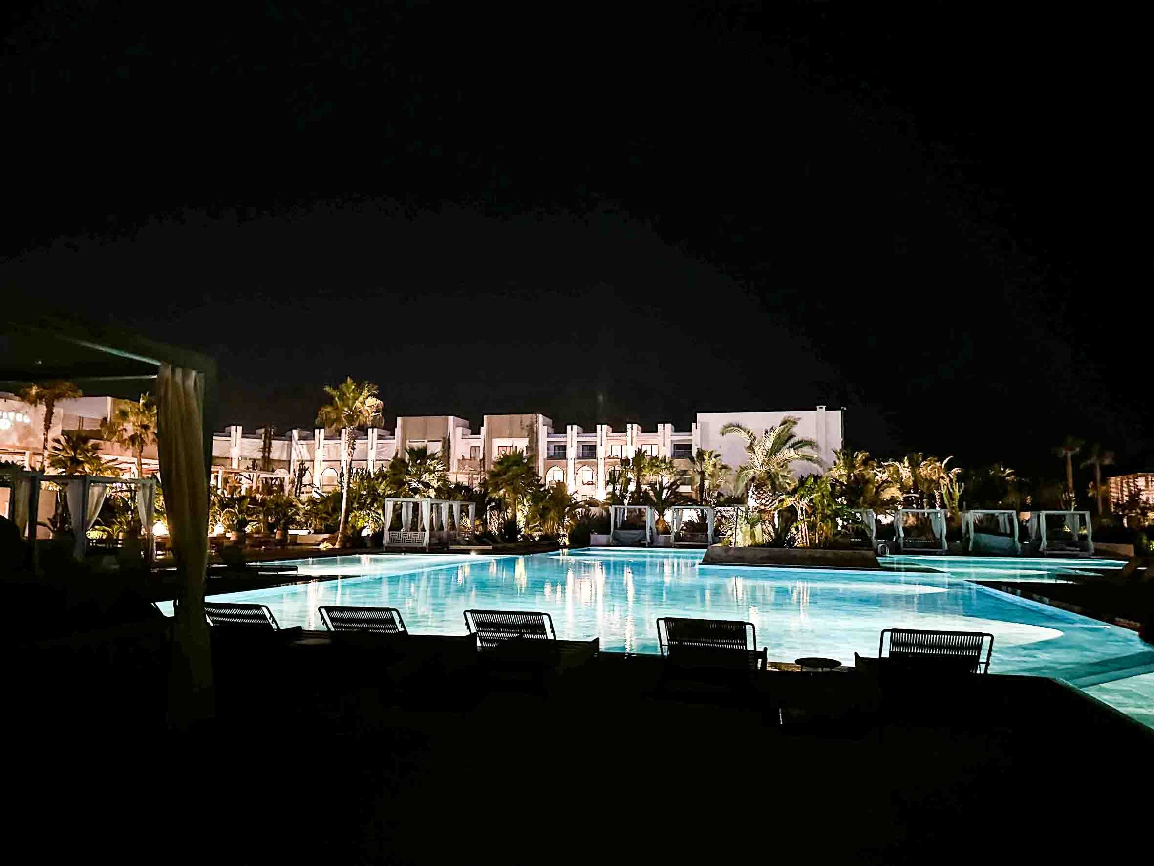 Night view of a hotel pool illuminated with blue lights, surrounded by palm trees and white cabanas, with a white building in the background.