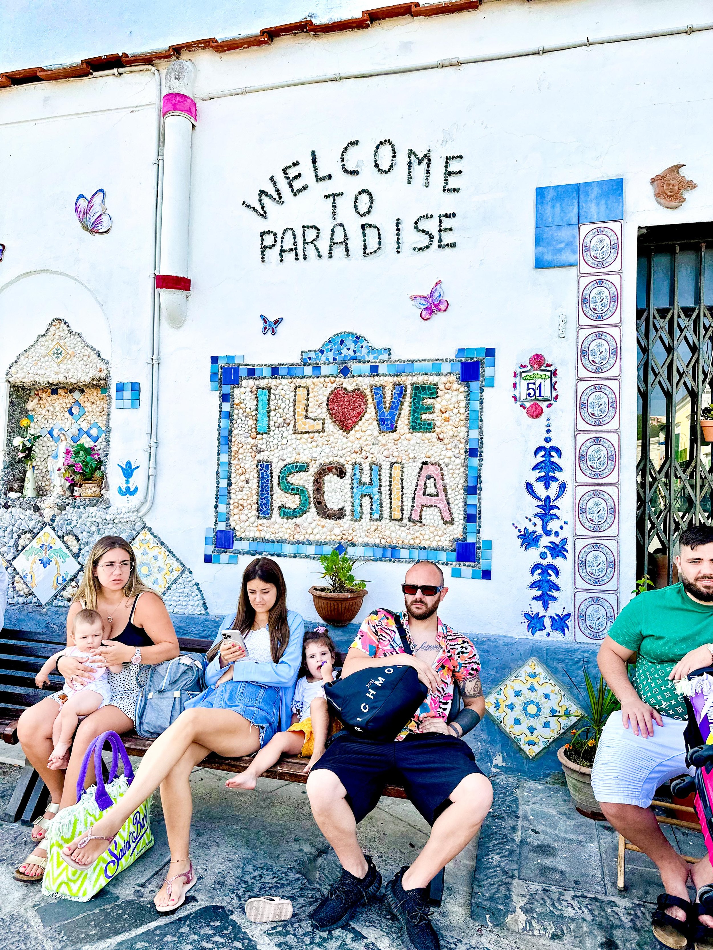 A group of five people sitting on benches in front of a colorful, decorative wall that says "Welcome to Paradise" and "I Love Ischia." The wall has mosaic and tile decorations with butterflies and flowers.