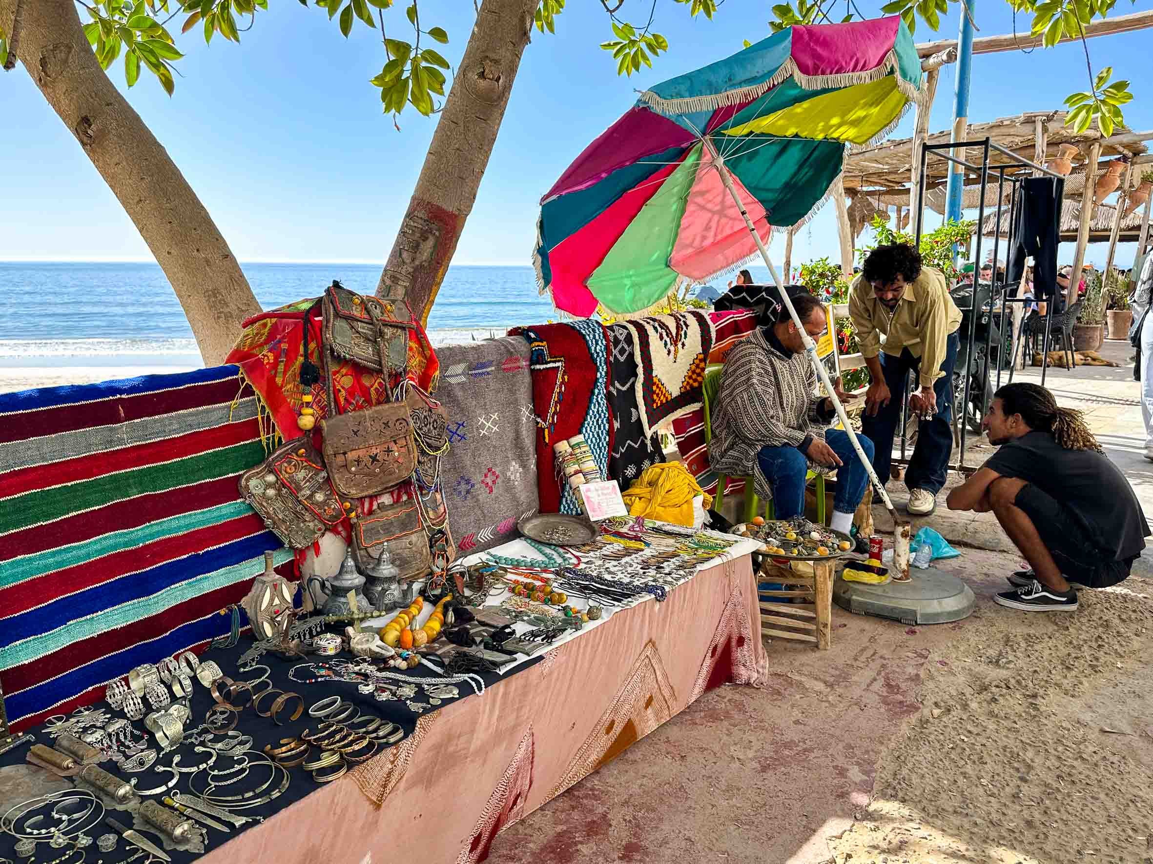 A beachside outdoor market with handmade jewelry, bags, and textiles displayed on tables and hung on a red, green, and blue striped cloth. Two people are seated under a large colorful umbrella, talking, with a man crouching in front of a woman, possi