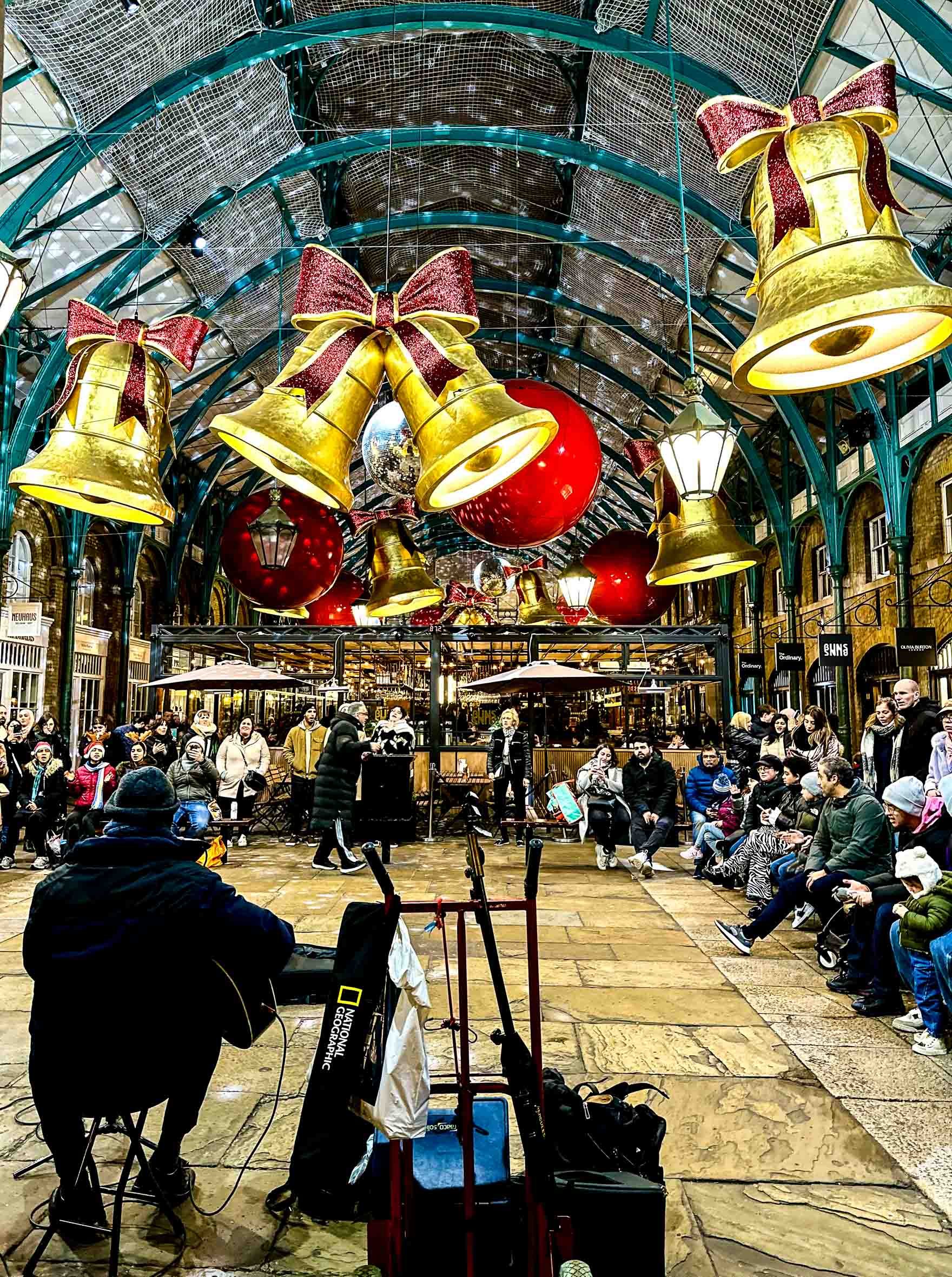 Indoor festive scene with hanging gold bells with red bows, red and gold baubles, and people gathered for a performance in a covered market or arcade.