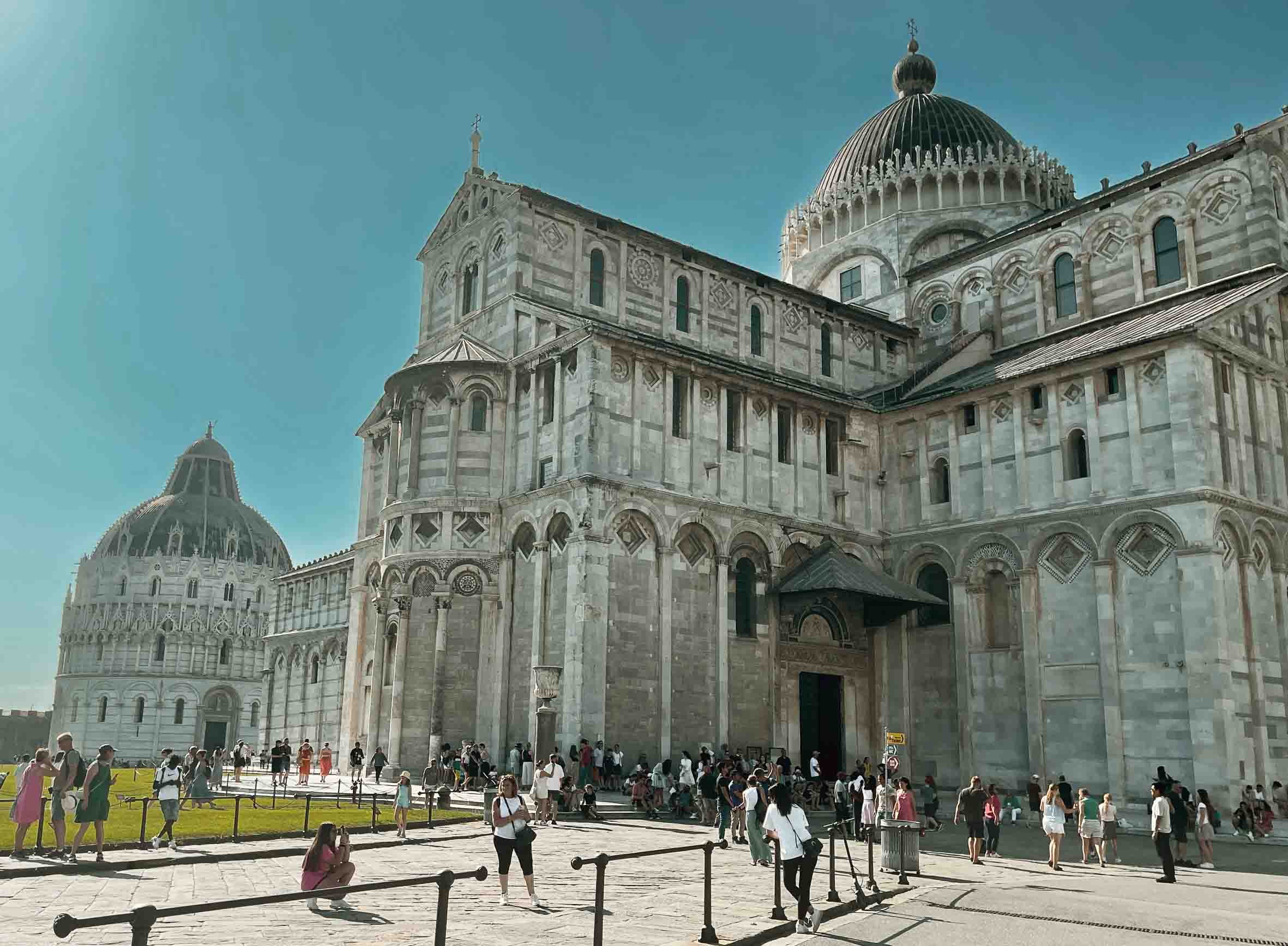 People gathering outside the Pisa Cathedral in Italy, a large historic church with ornate stonework and multiple domes under a clear sky.