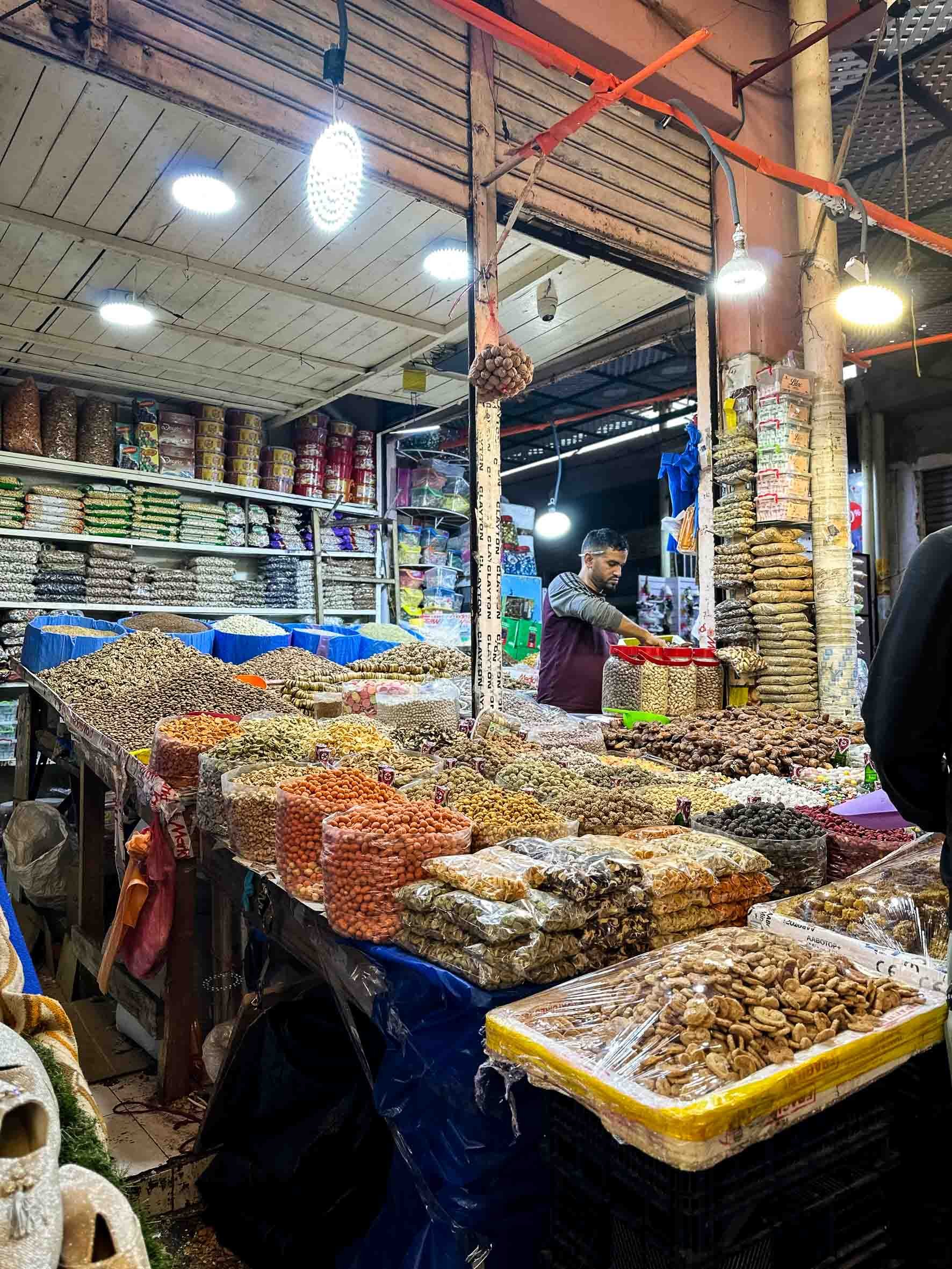 A market stall selling various dried nuts and legumes, with a man behind the counter and shelves filled with packaged goods in the background.