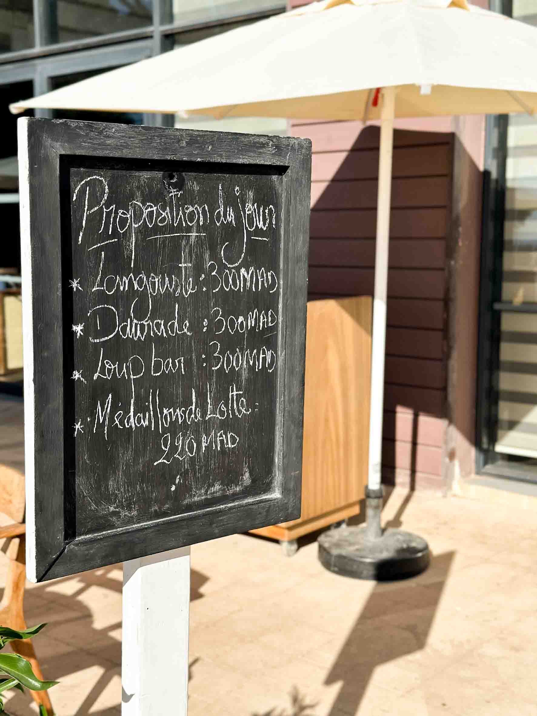 Outdoor menu board with handwritten listing of food options in front of a patio umbrella and building