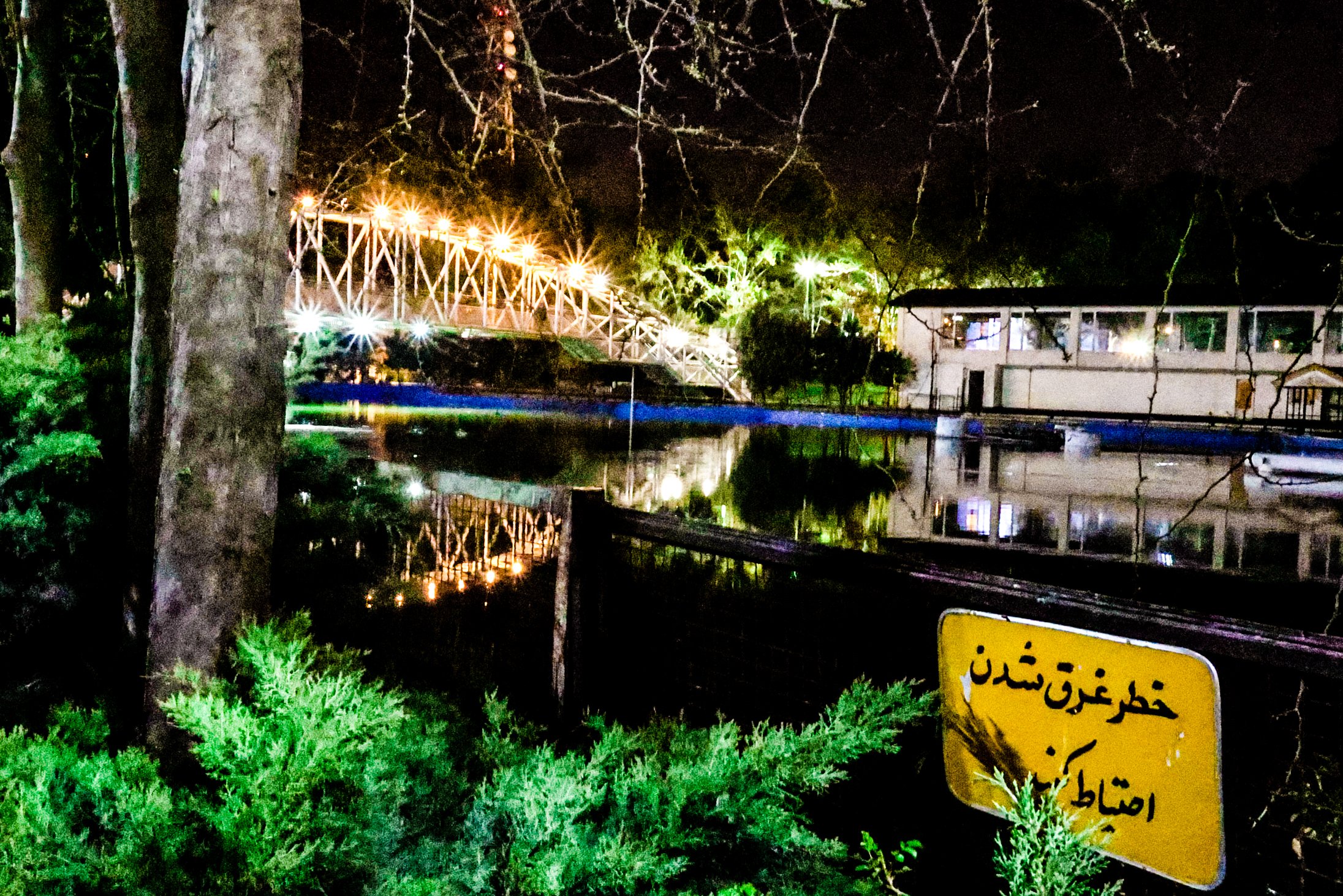 Nighttime scene of a well-lit bridge over a river, with their reflections visible in the water. In the foreground, there are green bushes and a yellow warning sign with Persian writing.