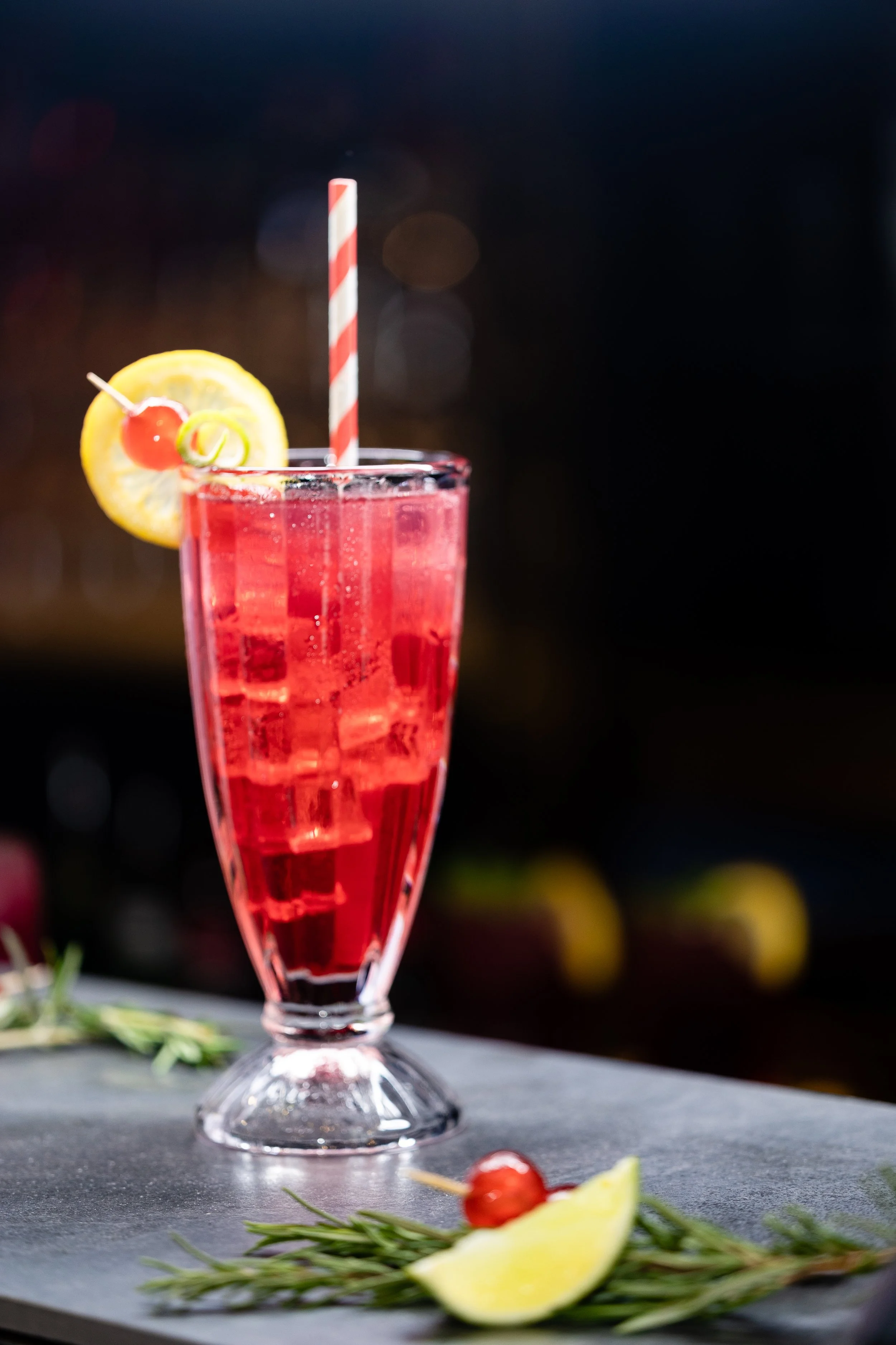 A tall glass of red cocktail with ice, garnished with a lemon slice and cherry, and a striped straw, placed on a gray surface with lemon wedges and rosemary sprigs in the foreground.