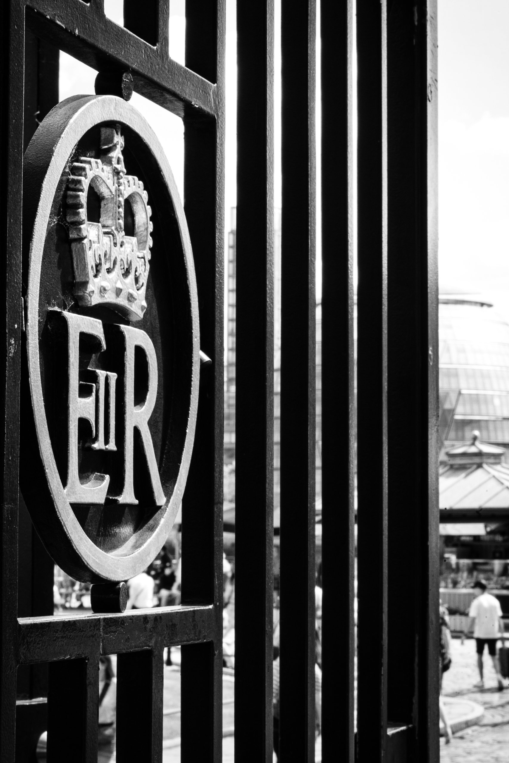 Close-up of the word 'FER' on a decorative iron gate with a crown emblem above, with a blurred background of people and a market or fair scene.
