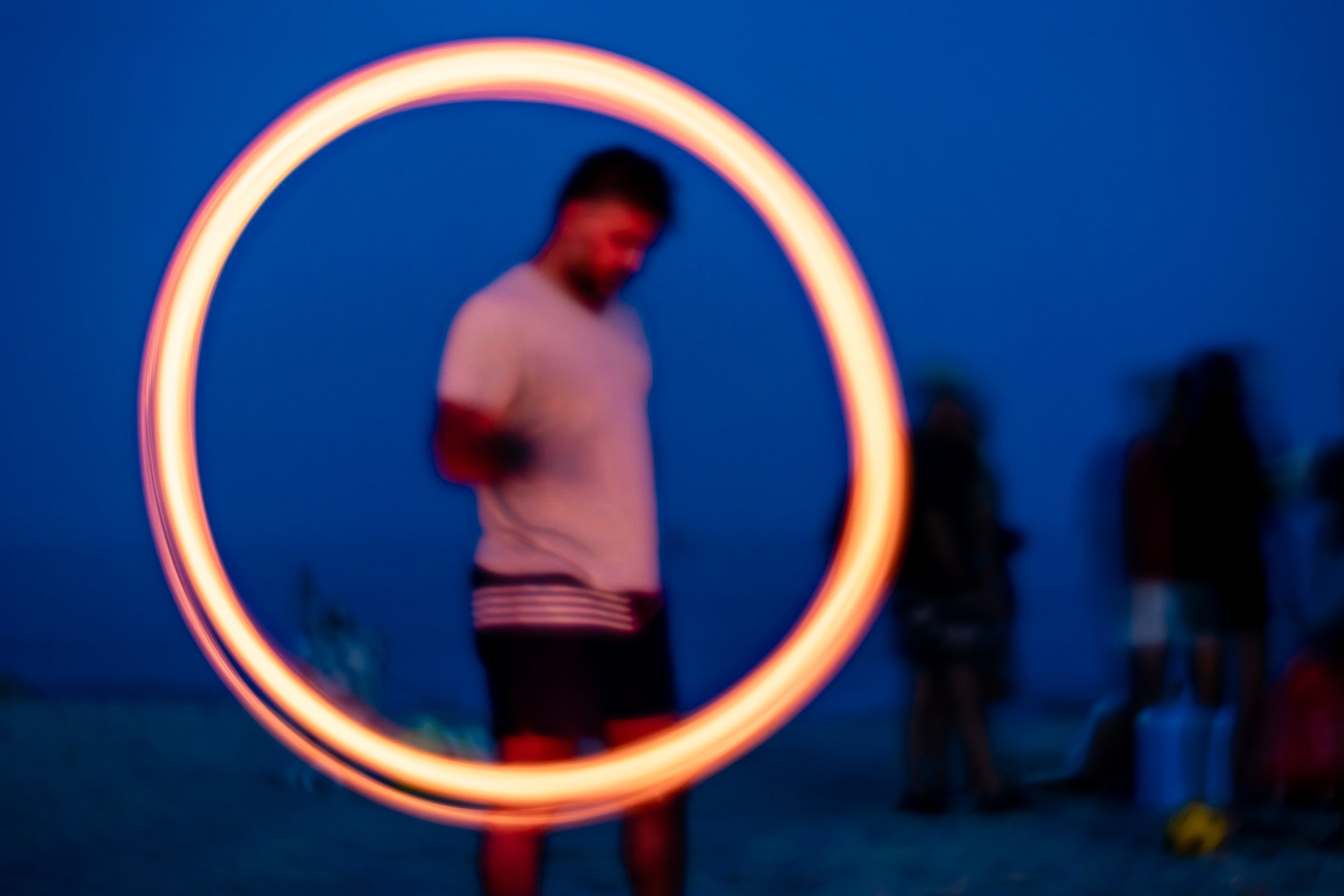 A person standing inside a glowing hula hoop during dusk at the beach, with blurred figures in the background.