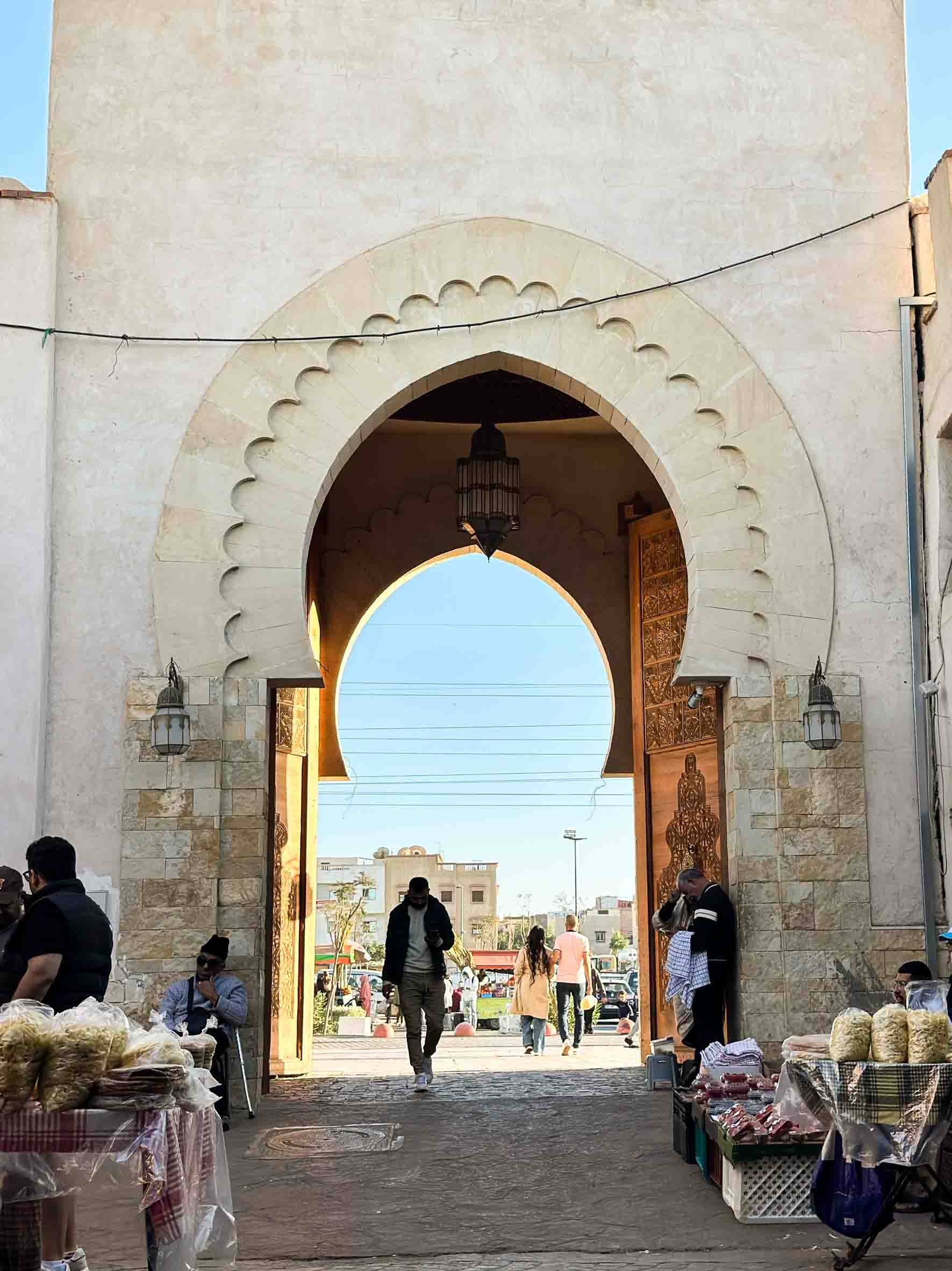 People walking and shopping through a large decorative archway, with market stalls selling various goods on either side.