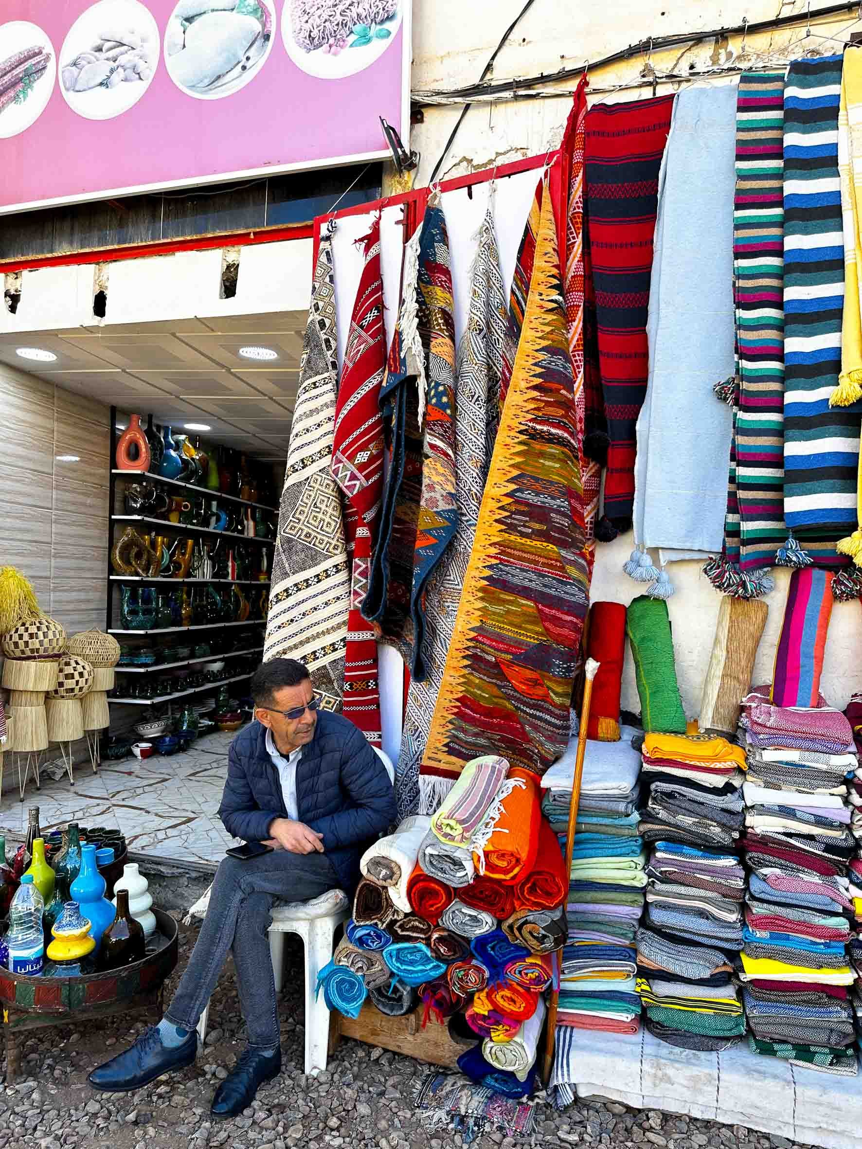 A man with sunglasses sitting on a white plastic chair at a street market stall selling colorful textiles, rugs, and blankets outside a shop.