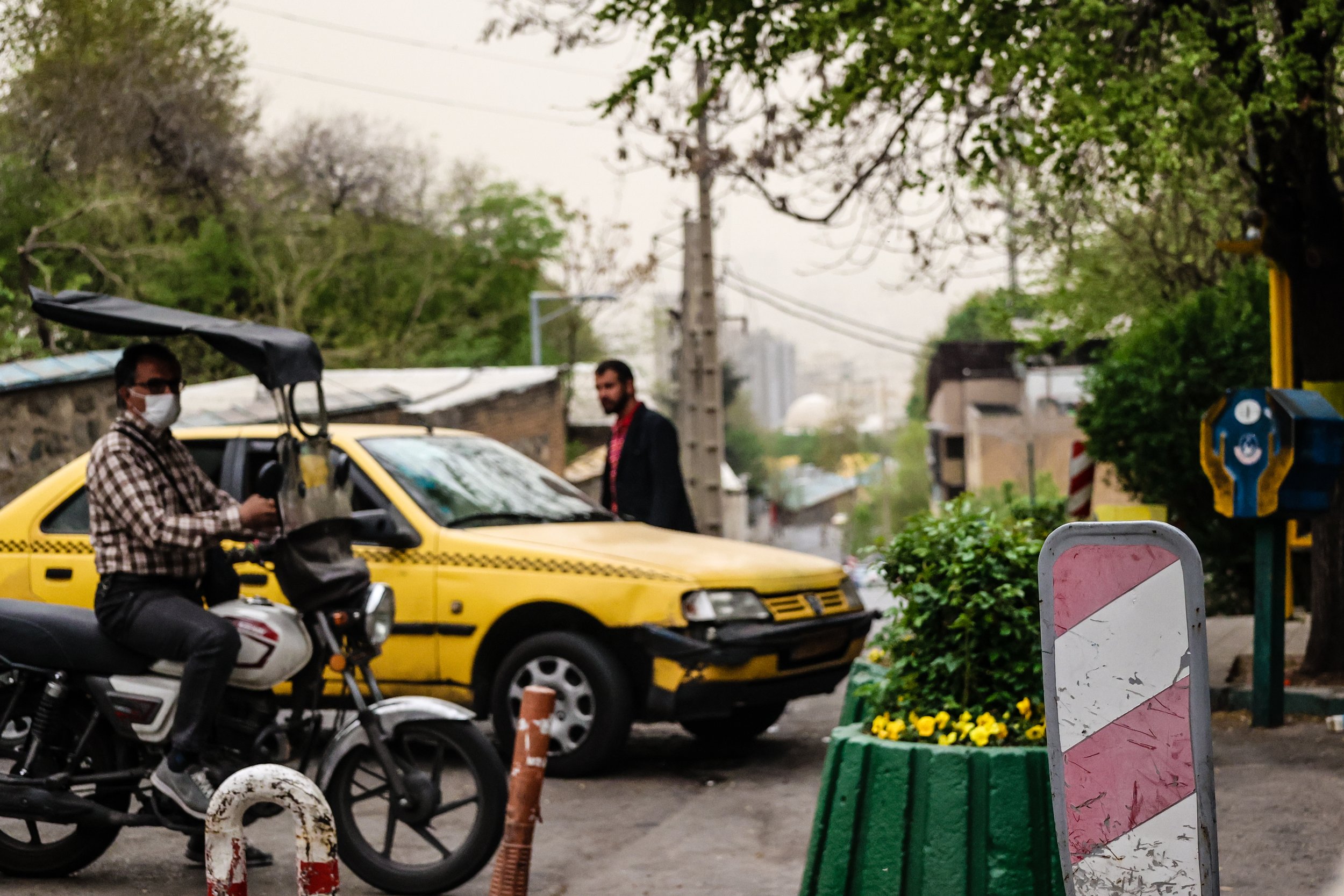 A man wearing a face mask and checkered shirt sitting on a motorcycle, a yellow taxi car in the background, and a person in a dark suit walking.