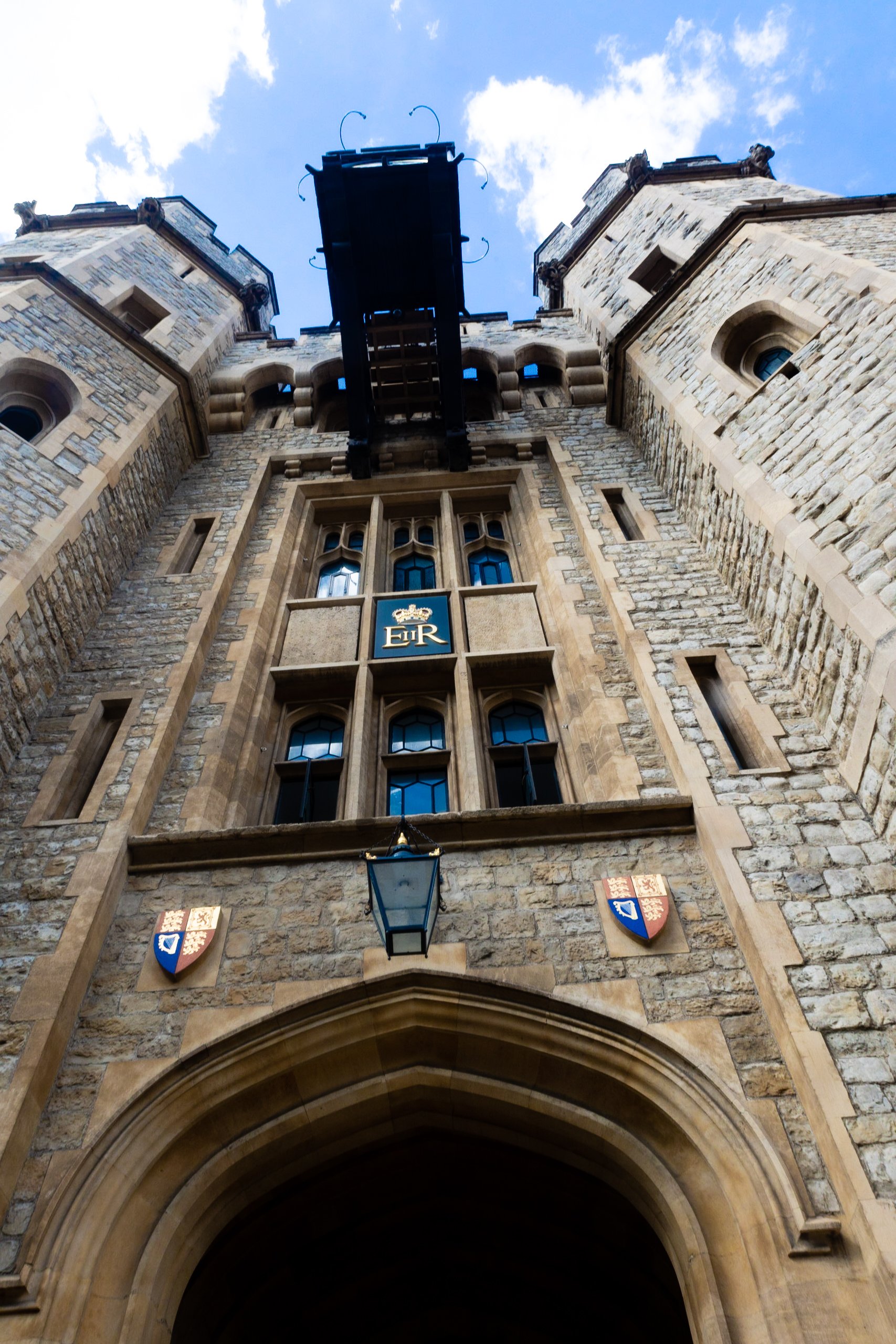 View from the ground looking up at a medieval castle tower with stone walls and a clock above an archway, featuring shields and a lamp hanging from the arch.