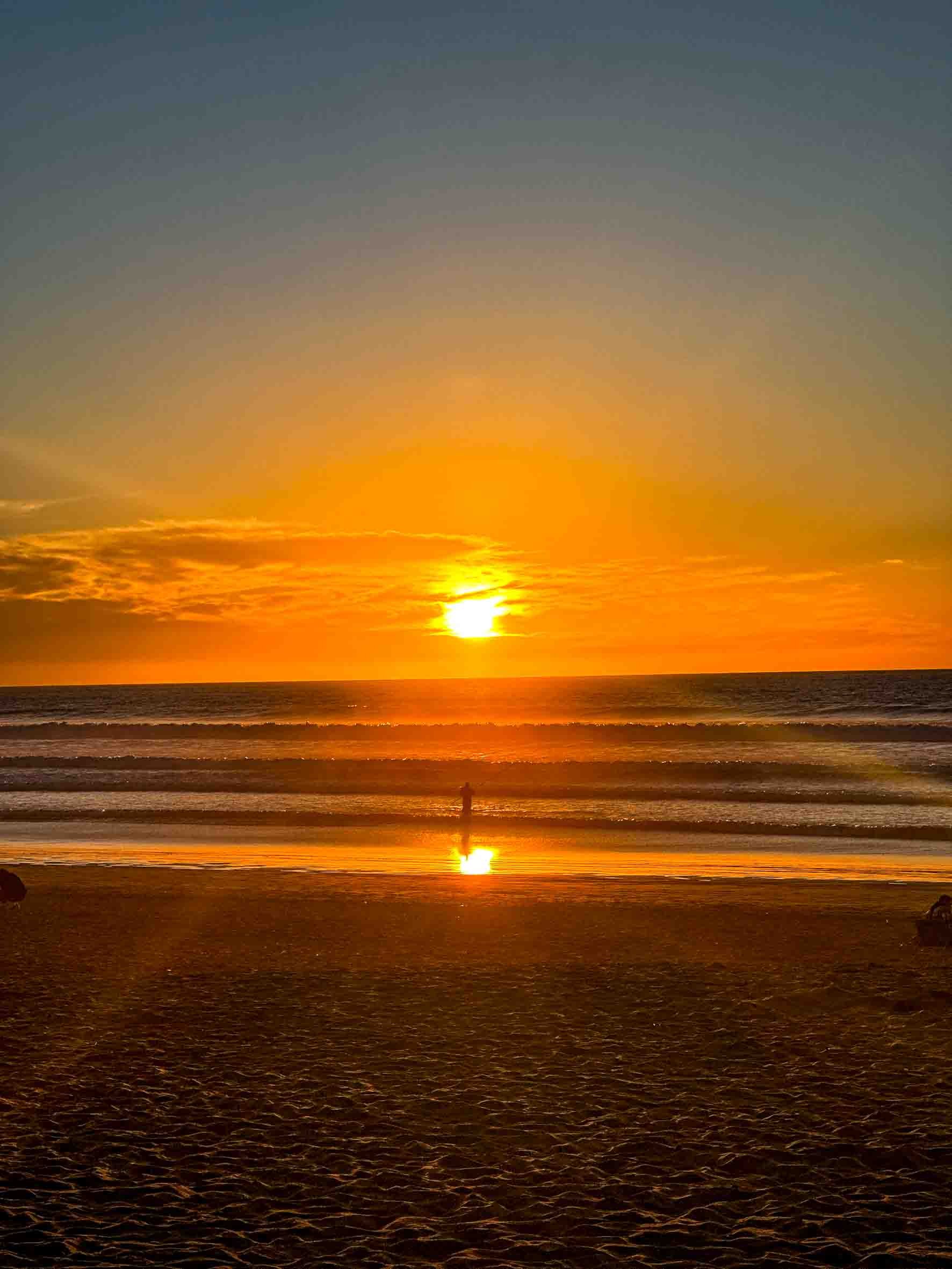 Sunset over the ocean with a person standing in the water, and the sky transitioning from orange near the horizon to dark blue at the top.
