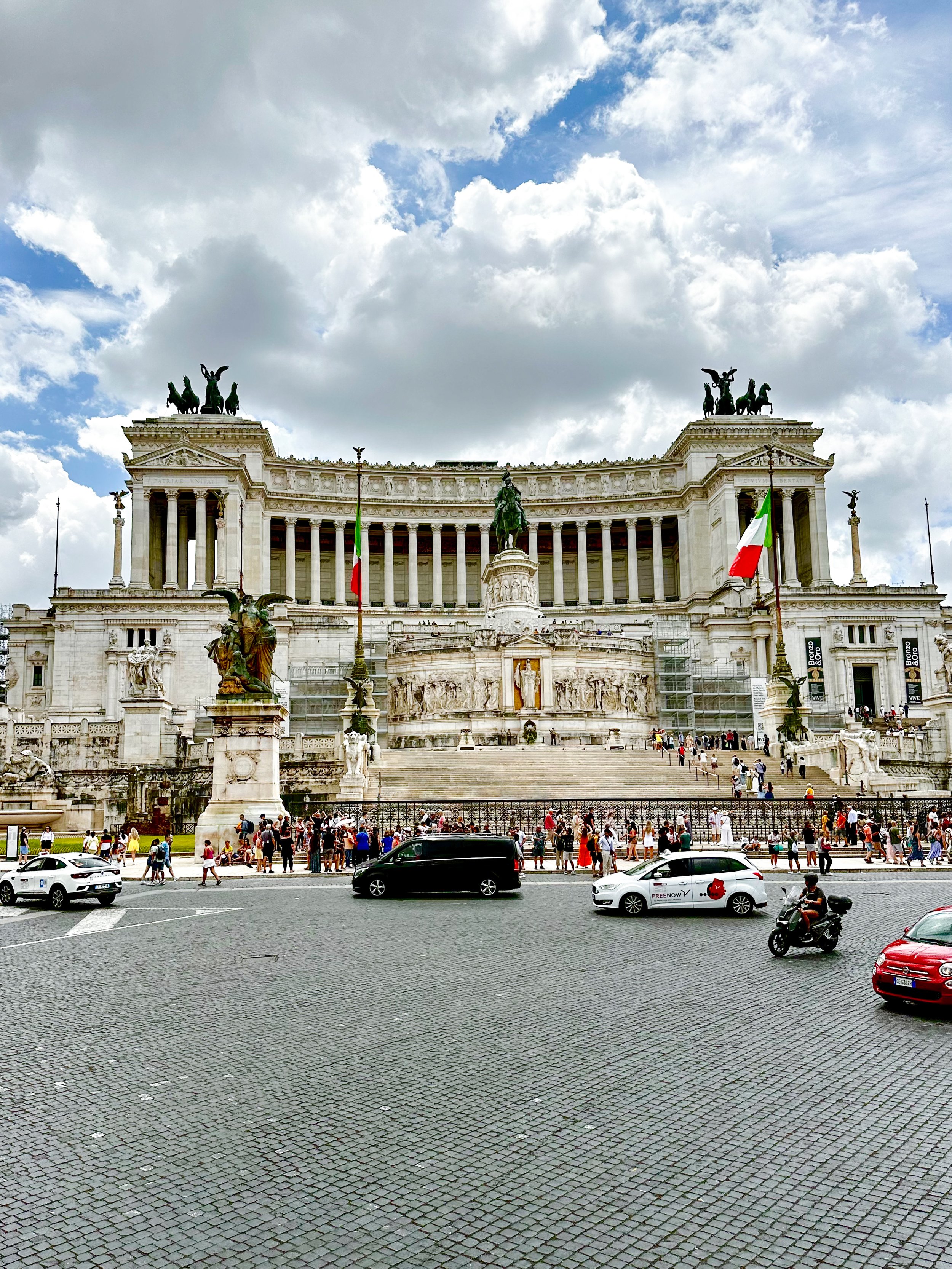 The Altare della Patria in Rome, Italy, a large white marble monument with statues and columns, Italian flags, and a crowd of visitors in front on a cloudy day.