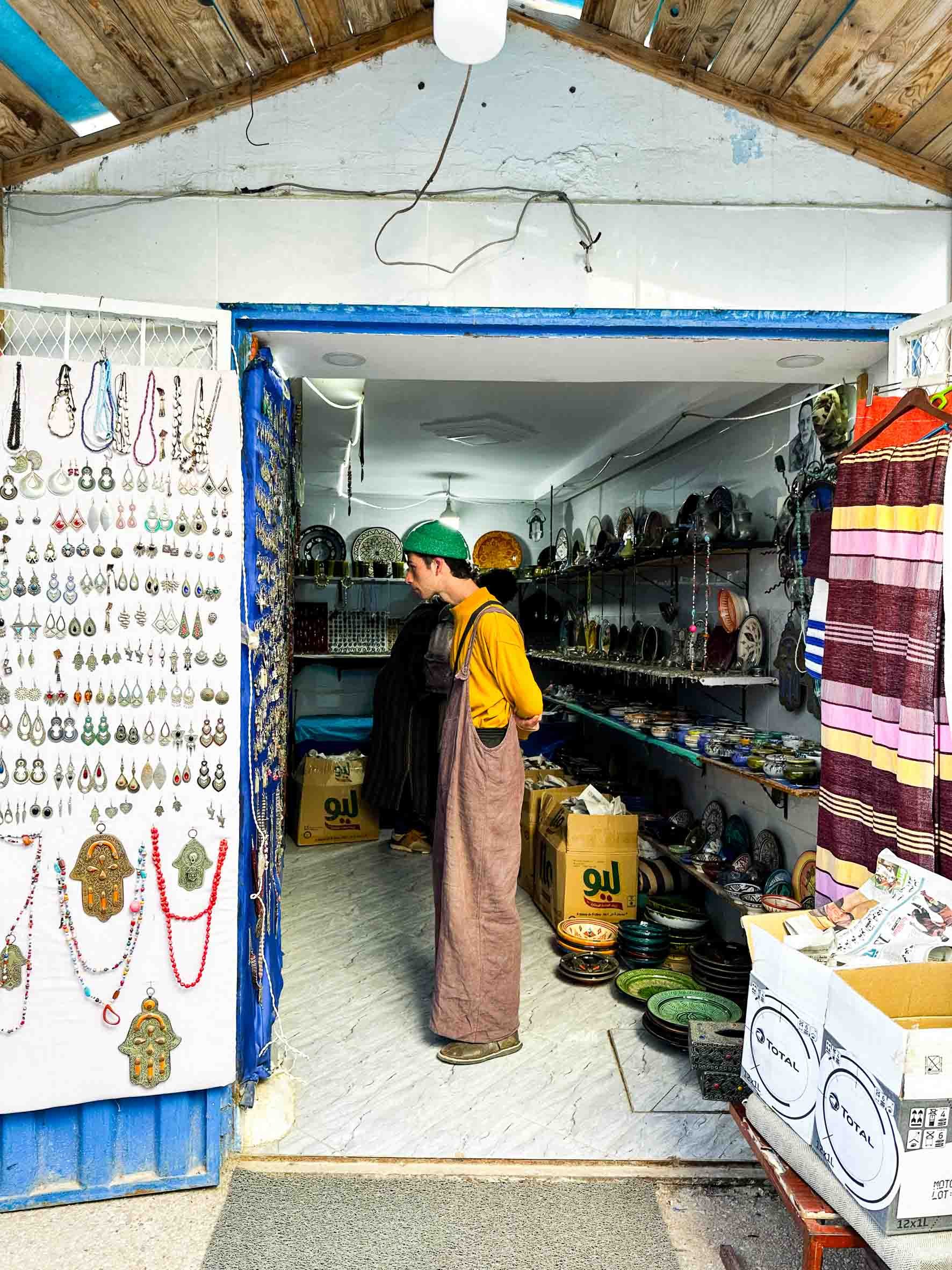 A person wearing a yellow shirt, a green cap, and beige pants shopping at a market stall with jewelry and pottery for sale. The stall has shelves filled with ceramics and a display of earrings, necklaces, and other jewelry on a white display board. T
