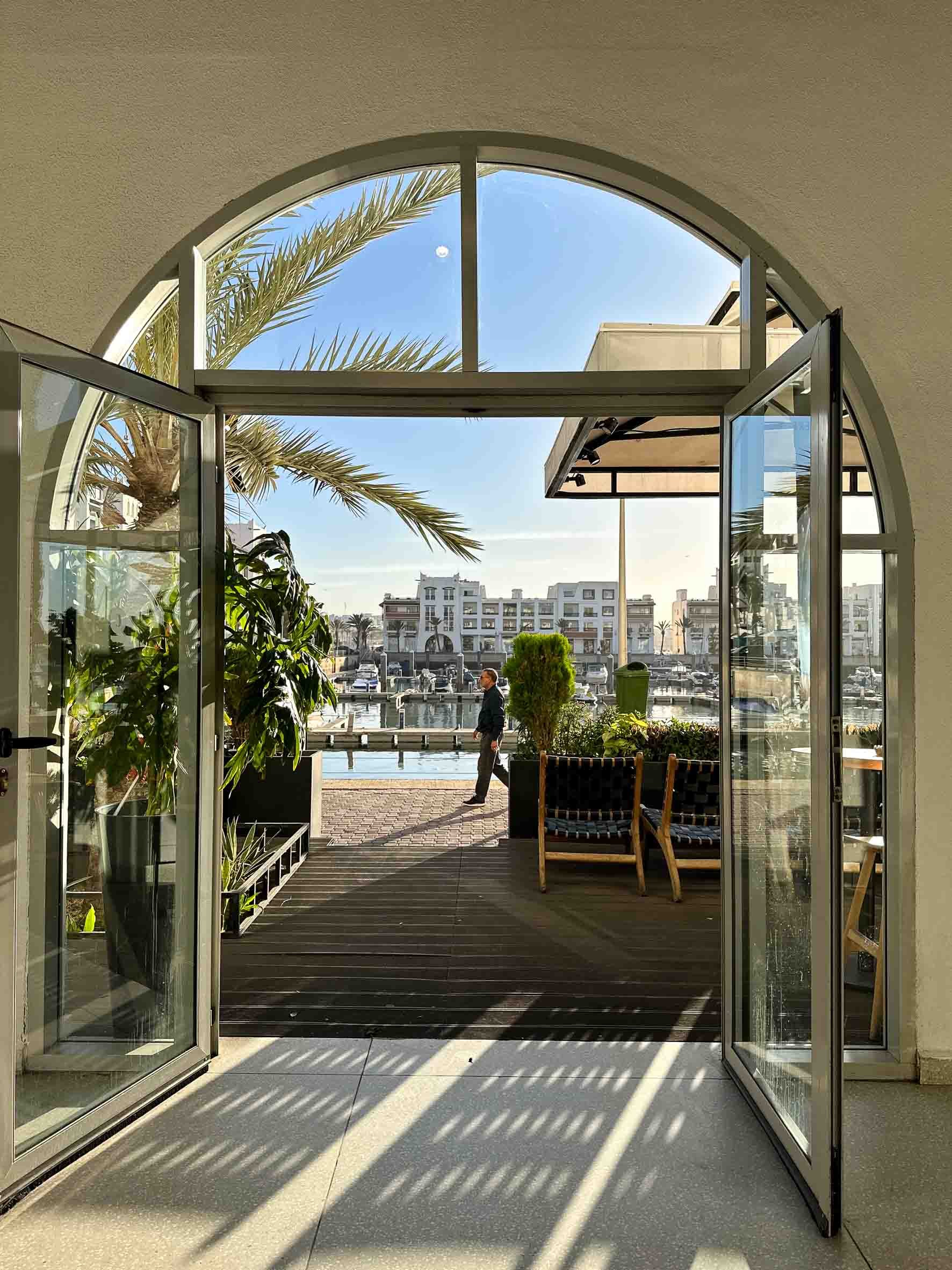 Opening double glass doors leading to a marina with boats and apartment buildings in the background. There are potted plants and outdoor seating on a wooden deck, sunny weather, blue sky with a visible moon.