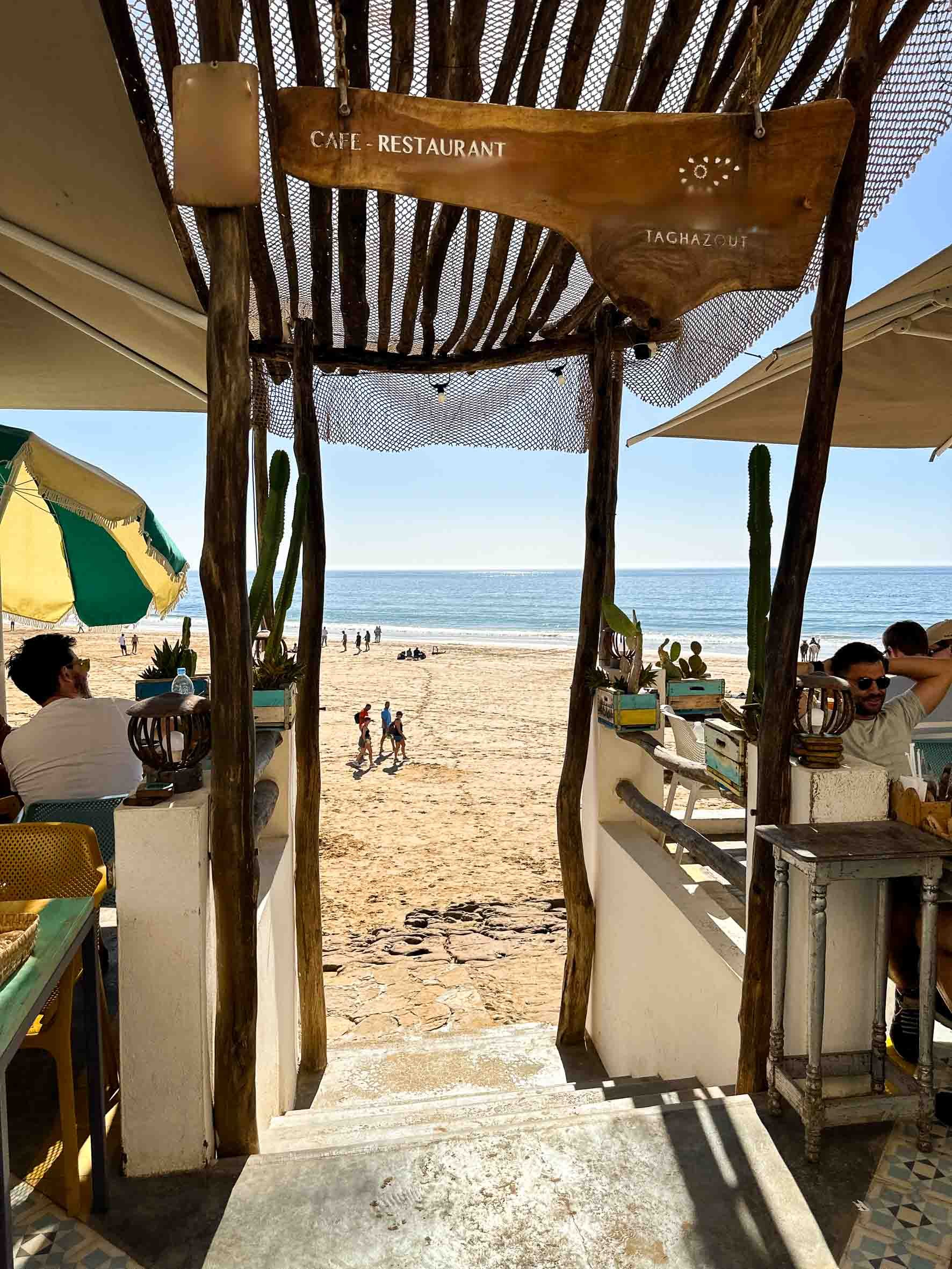 Beach view through entrance of a seaside cafe with wooden sign overhead pointing to the cafe and takeaway options, sandy beach with people walking and relaxing, bright sunny day.
