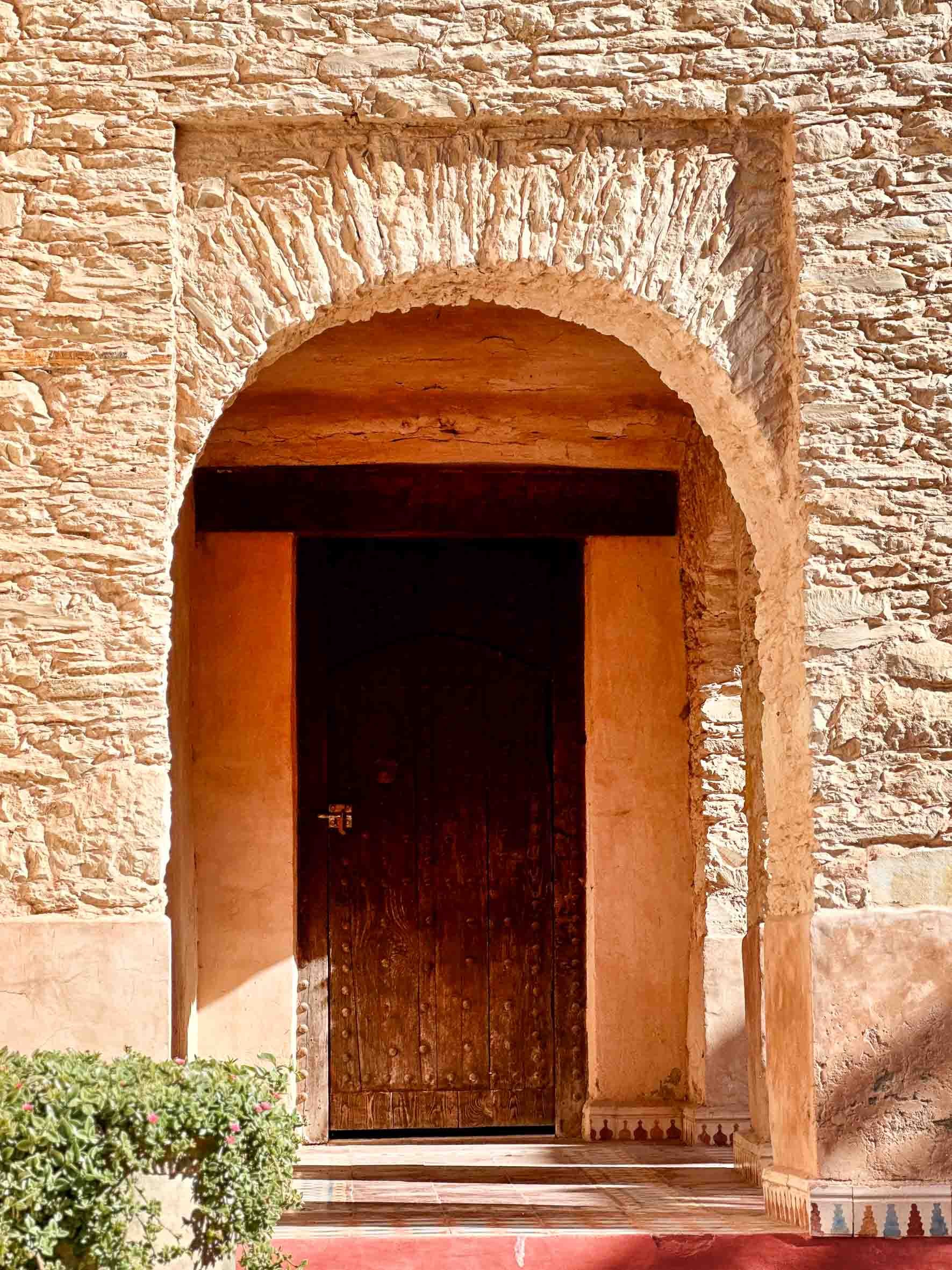 A rustic wooden door set within an arched stone doorway, with stone walls and a small bush in front.