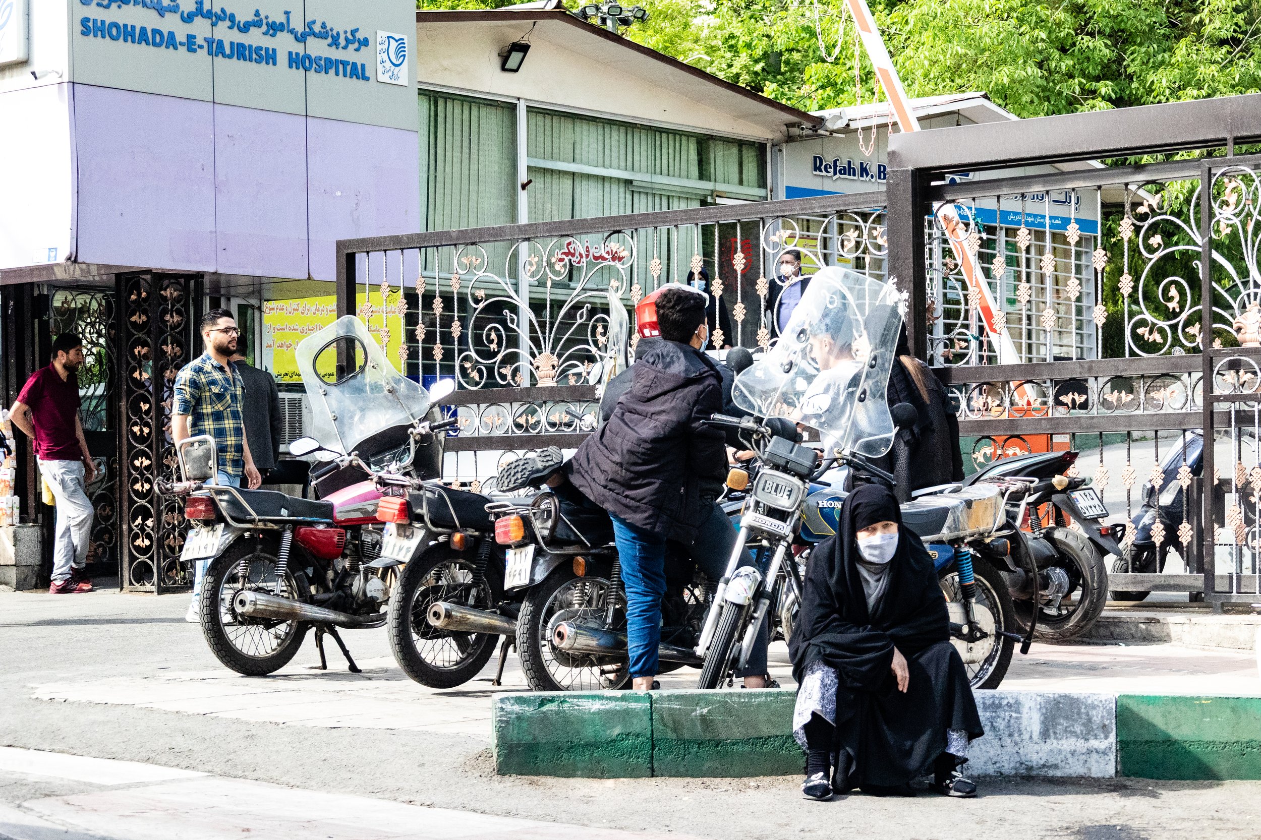 People waiting outside Shohada-e-Tajrish Hospital, some wearing masks, with motorcycles parked nearby and a woman seated on the sidewalk.
