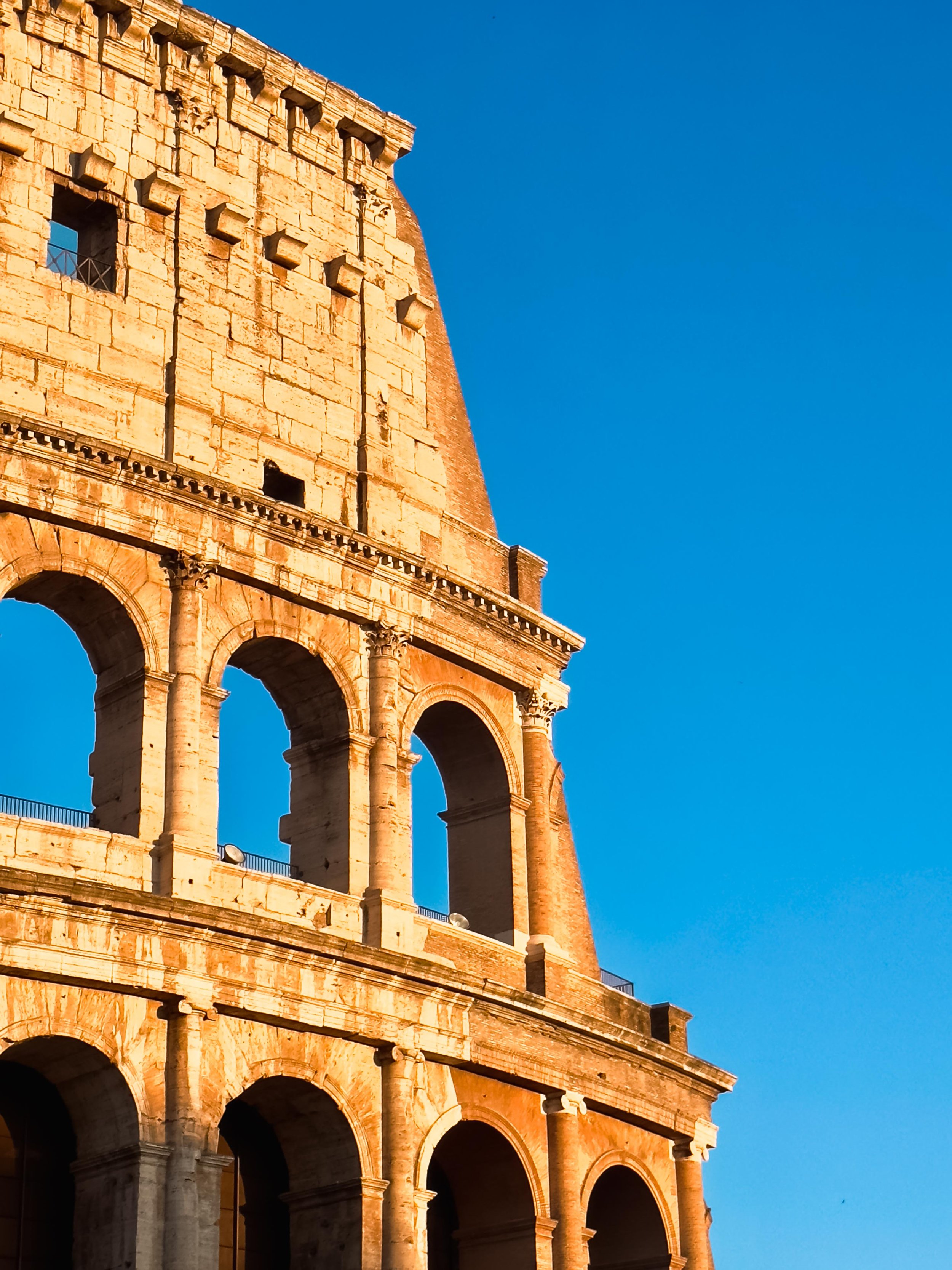 Close-up photo of the ancient Roman Colosseum's exterior with arches and stonework against a clear blue sky.