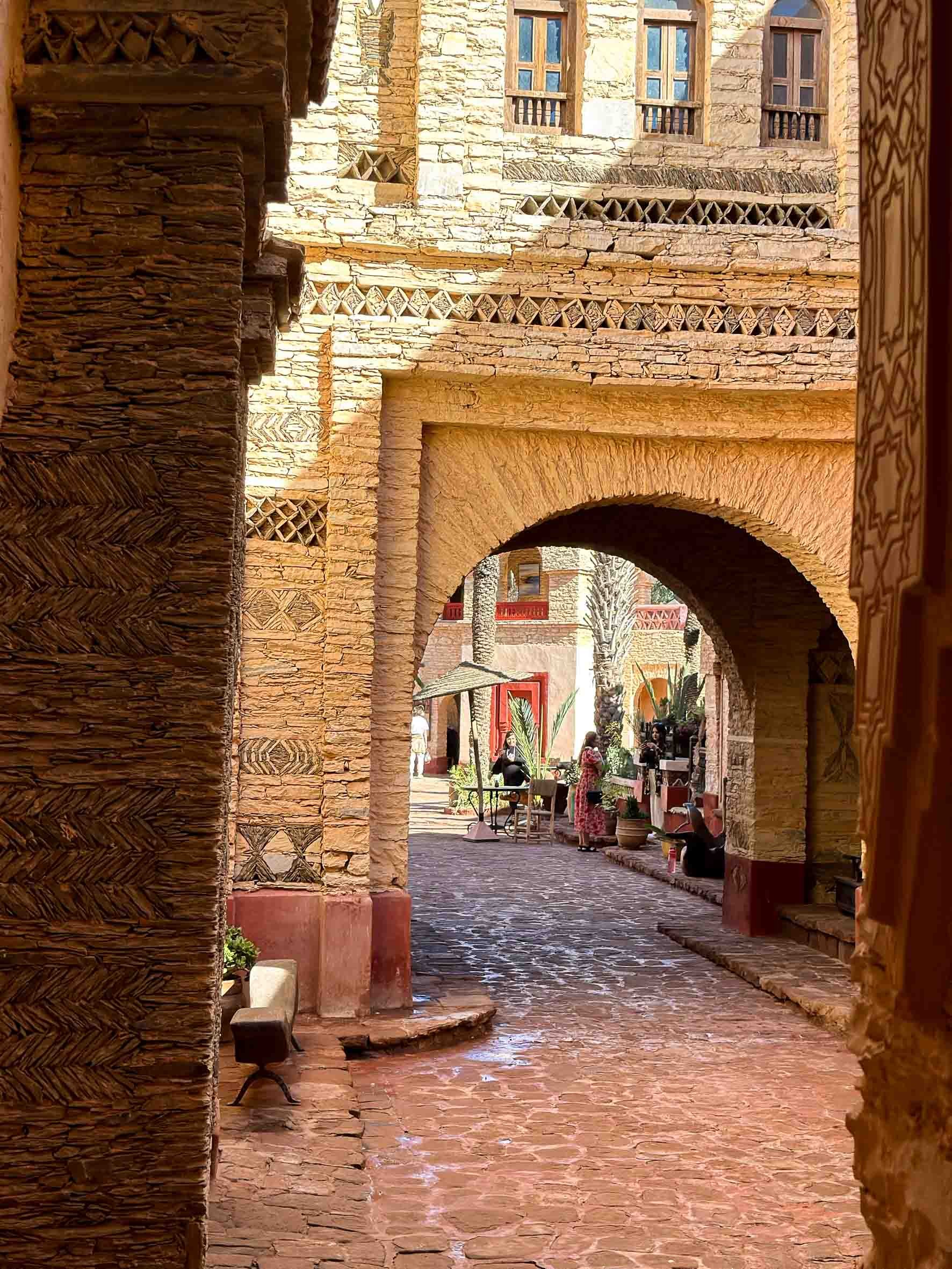 A narrow, cobblestone alleyway in a Mediterranean-style village with arches, stone walls, potted plants, and people sitting at outdoor tables.