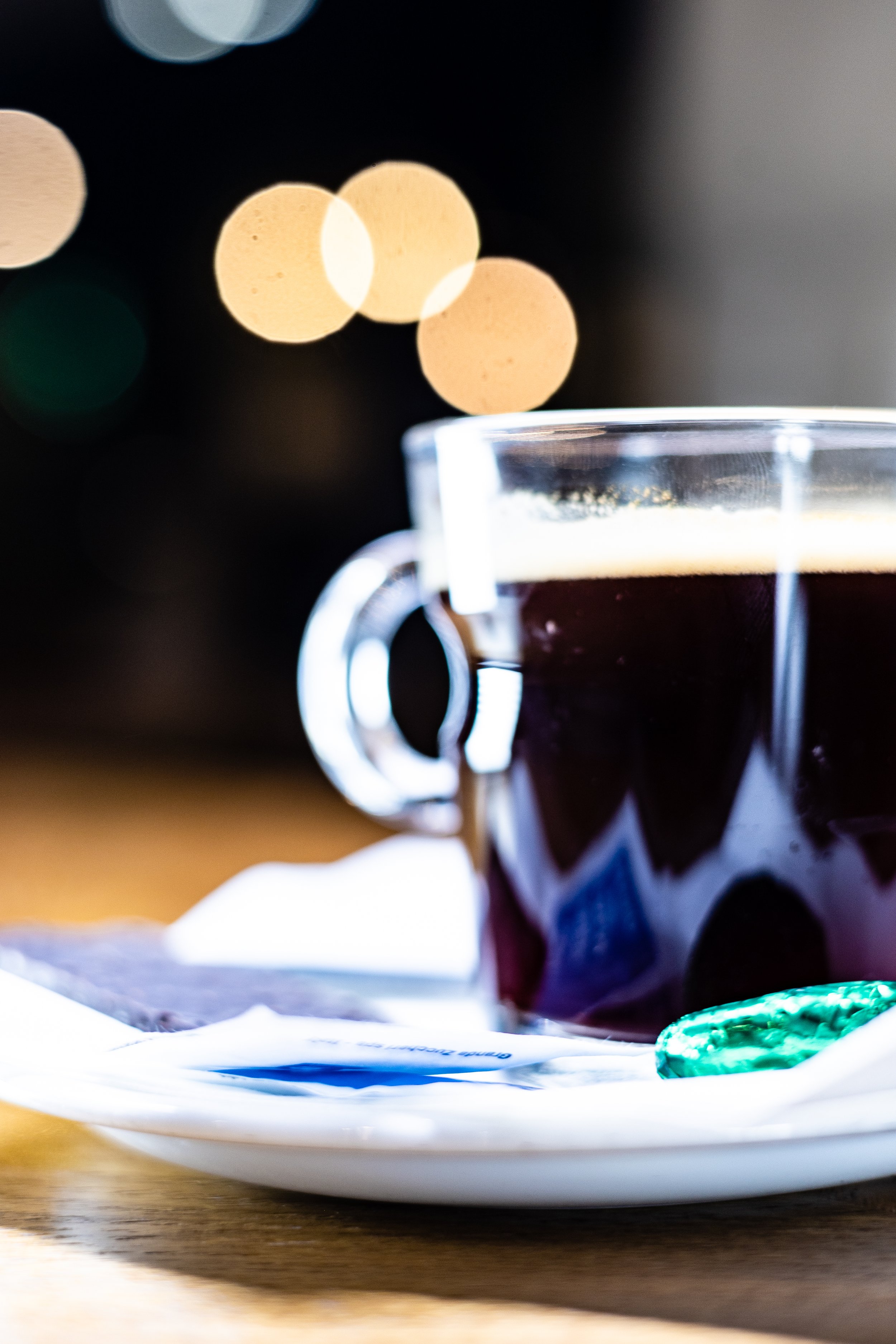 A clear glass cup filled with black coffee on a white saucer with a wrapped mint candy, on a wooden surface, with blurred colorful lights in the background.