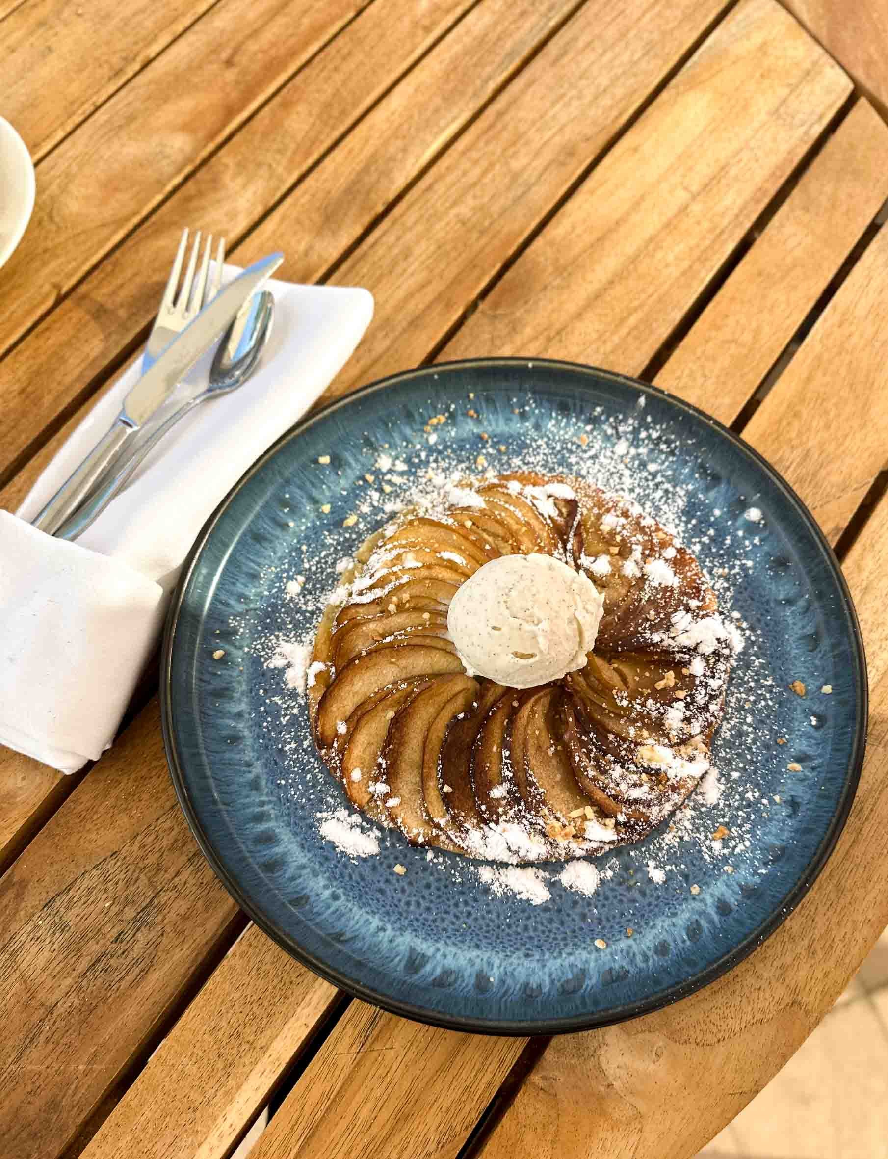 A plate of apple tarte with a scoop of ice cream on top, dusted with powdered sugar, on a blue ceramic plate on a wooden table.