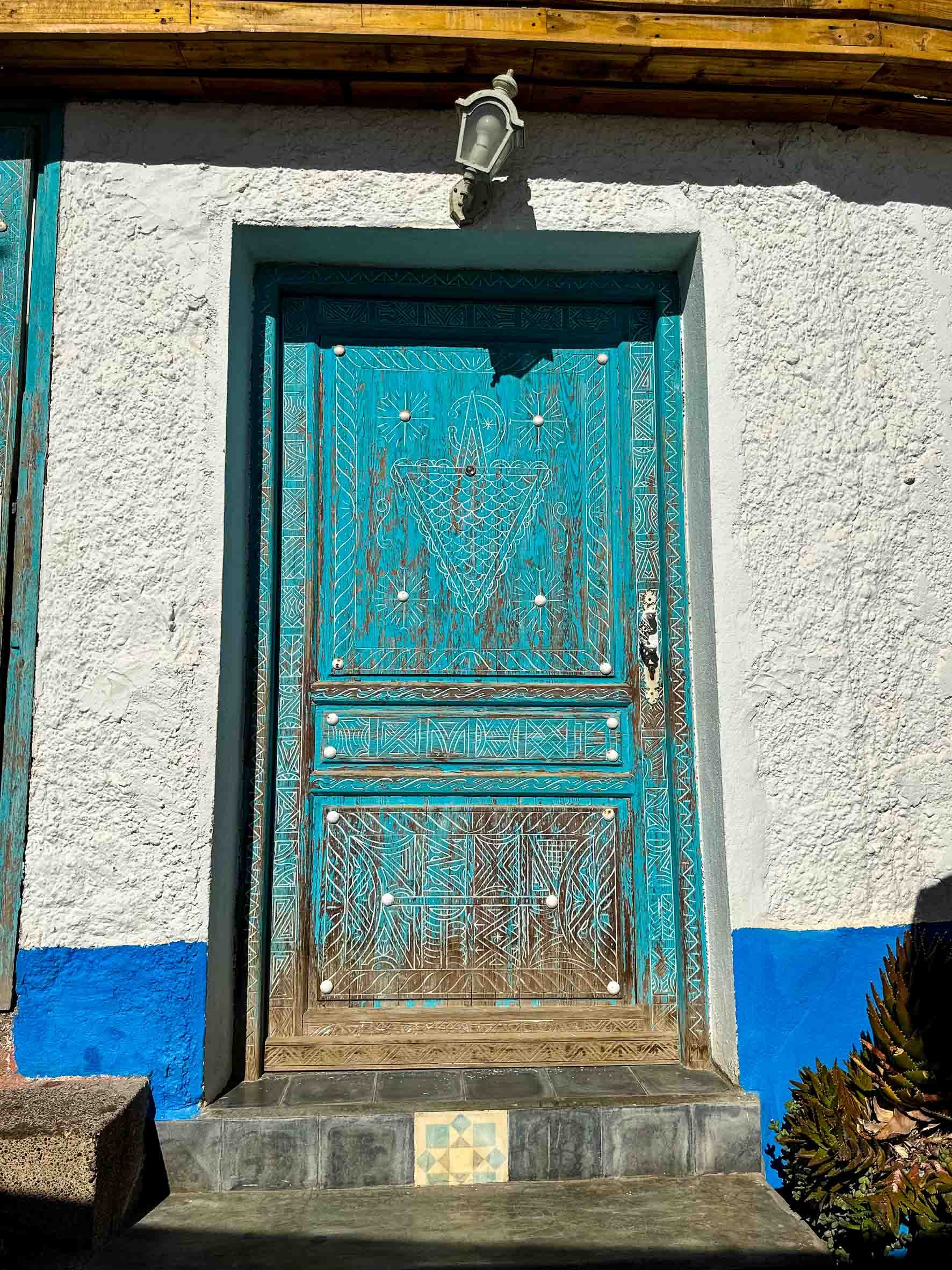 Decorative blue wooden door with geometric carvings, metal studs, and a small step in front, set into a white stucco wall with a blue painted section at the bottom, and an external lamp above.