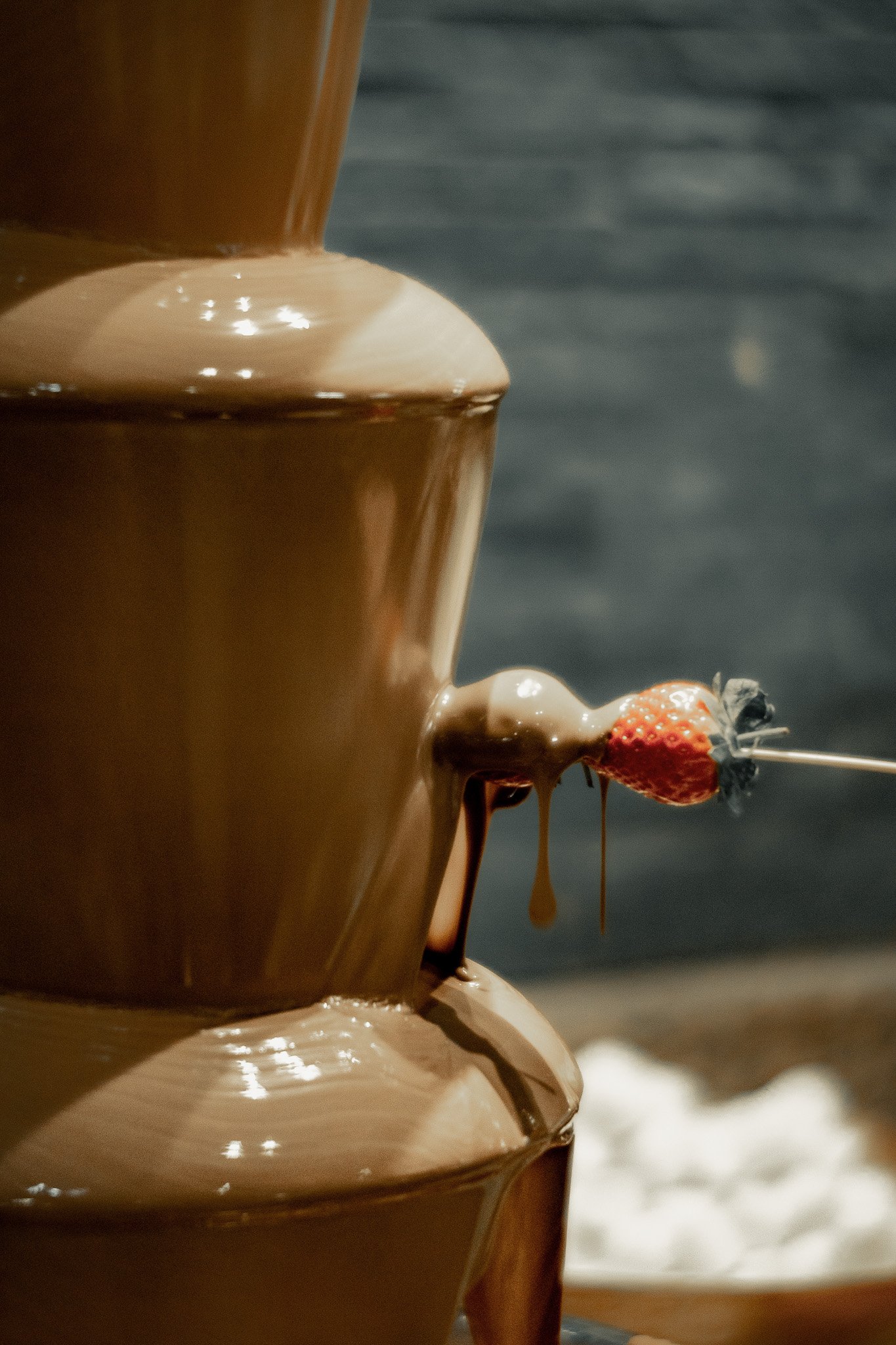 A chocolate fountain with melted chocolate flowing over tiers. A strawberry on a skewer is dipped in the chocolate fountain.