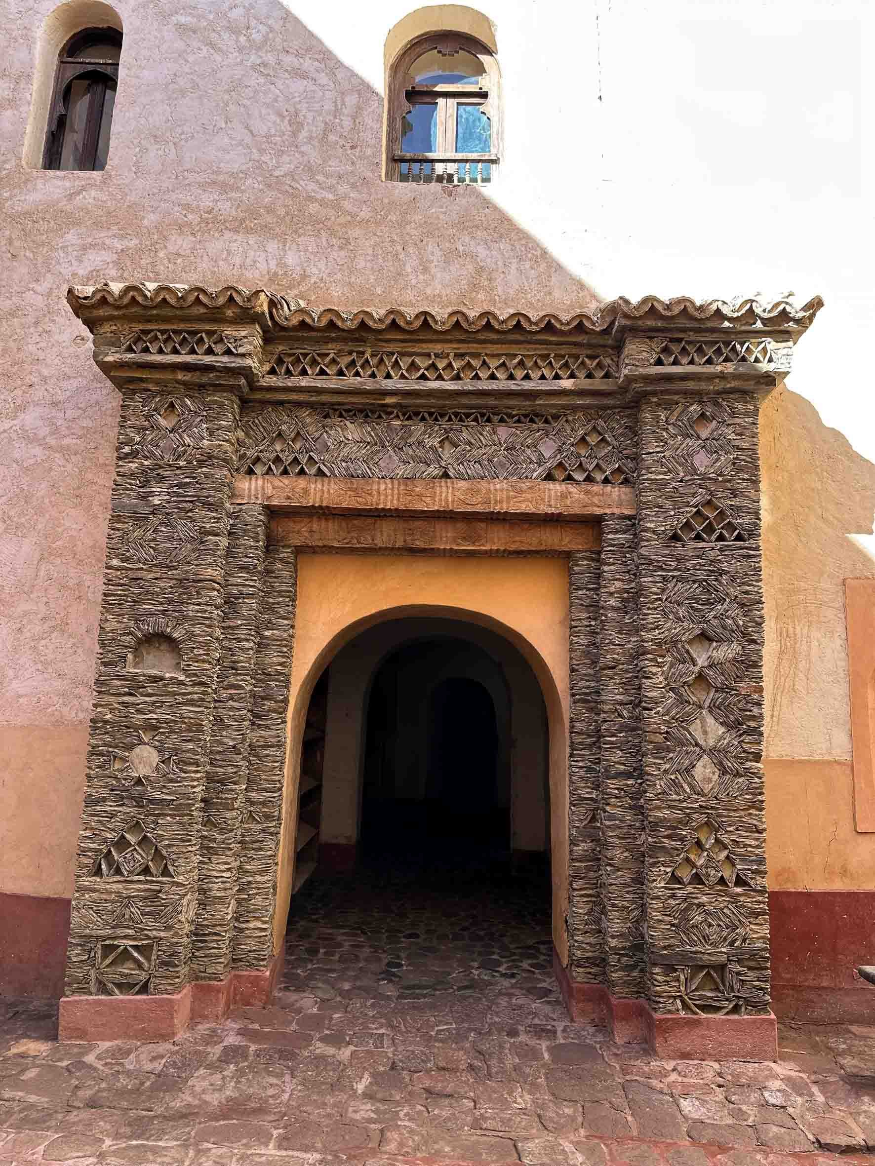 Stone archway entrance to a building with intricate carved wood and stone details, peach-colored walls, and a cobblestone pathway.