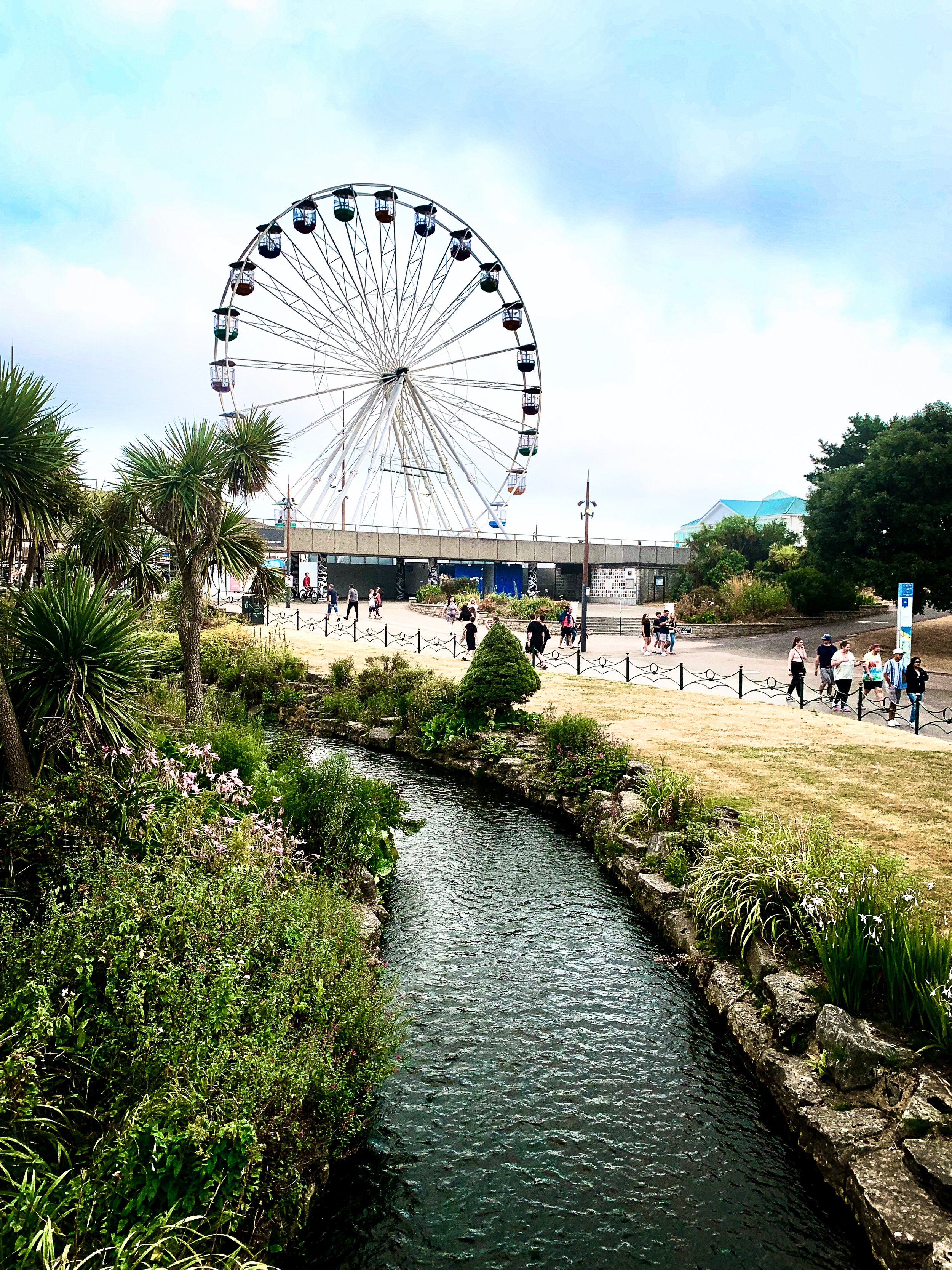 A Ferris wheel in an amusement park, with a river and lush greenery in the foreground, and people walking along a pathway.