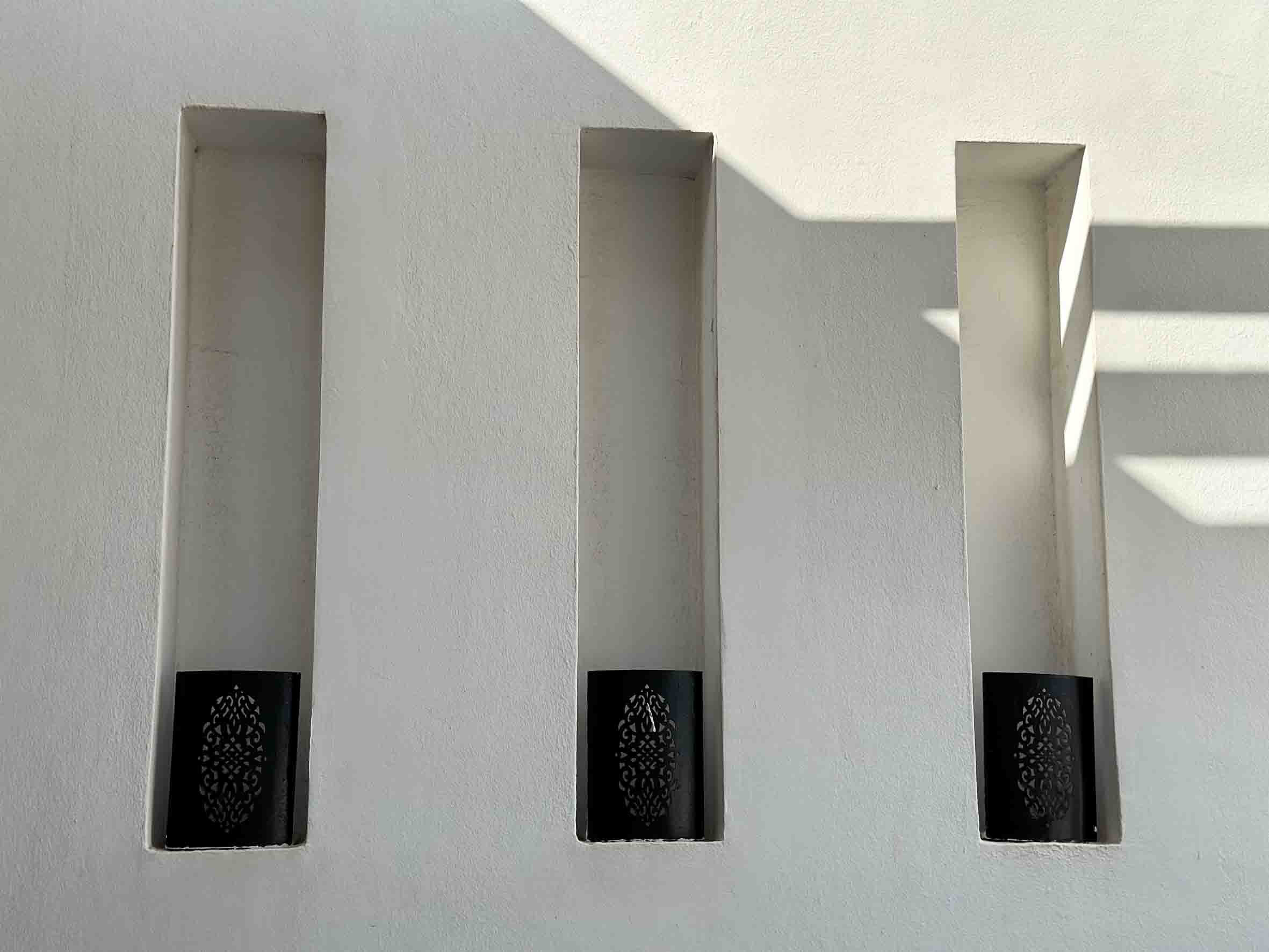 White wall with three vertical rectangular cutouts, each containing a black decorative element at the bottom, with shadows cast on the upper right side.