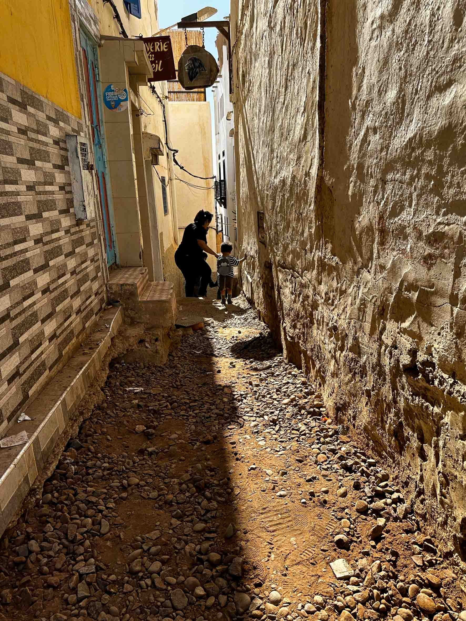 A narrow alleyway with uneven gravel and dirt ground, flanked by weathered stone and tile walls. A woman and a young child are walking together, with the woman helping the child. The background shows the upper parts of buildings with laundry hanging.