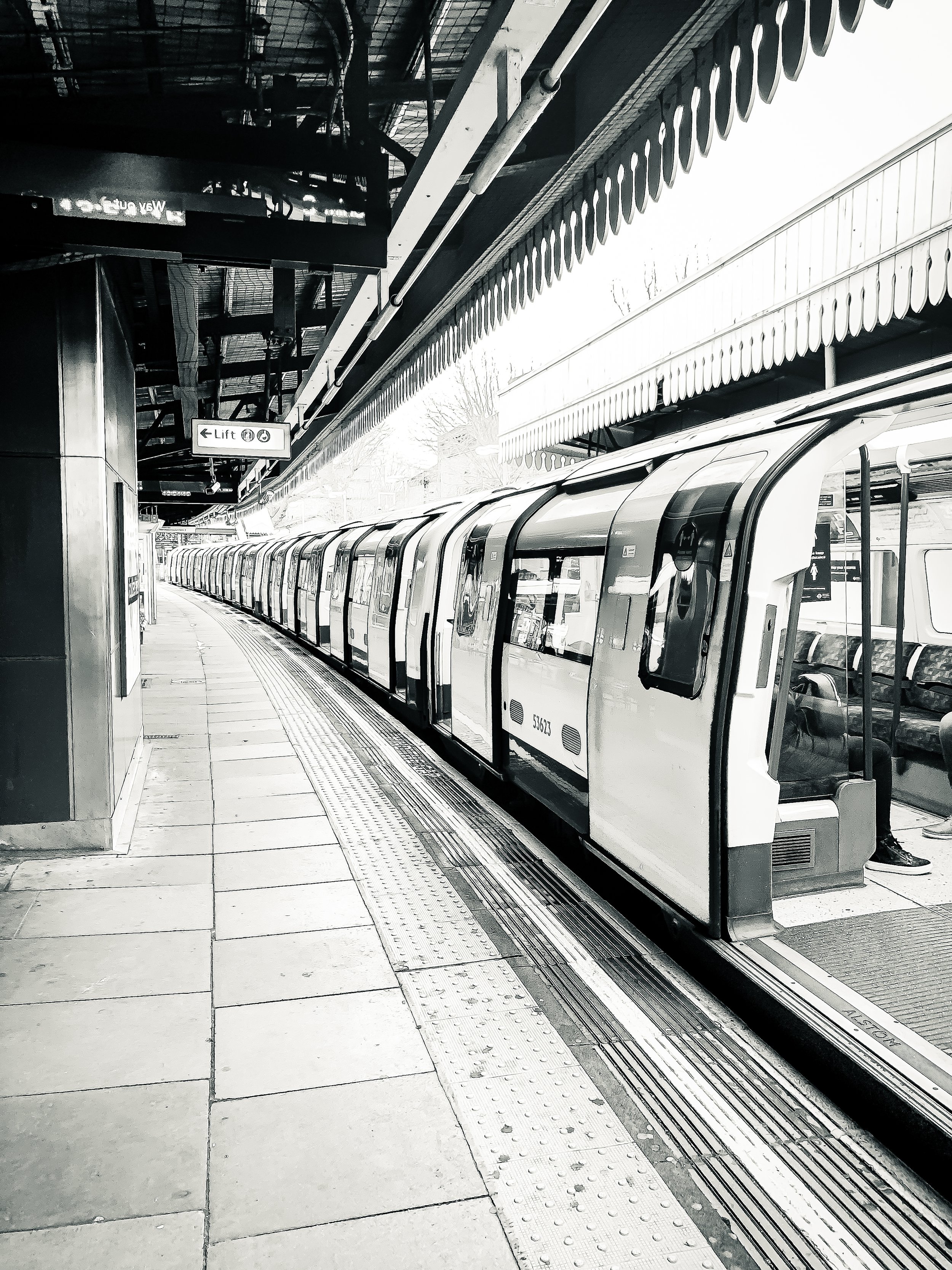A black and white photo of a modern train stopped at an outdoor station platform with a canopy overhead. The train has rounded doors and windows, and a sign indicating the lift is visible above. There is a sign hanging from the canopy showing the dir
