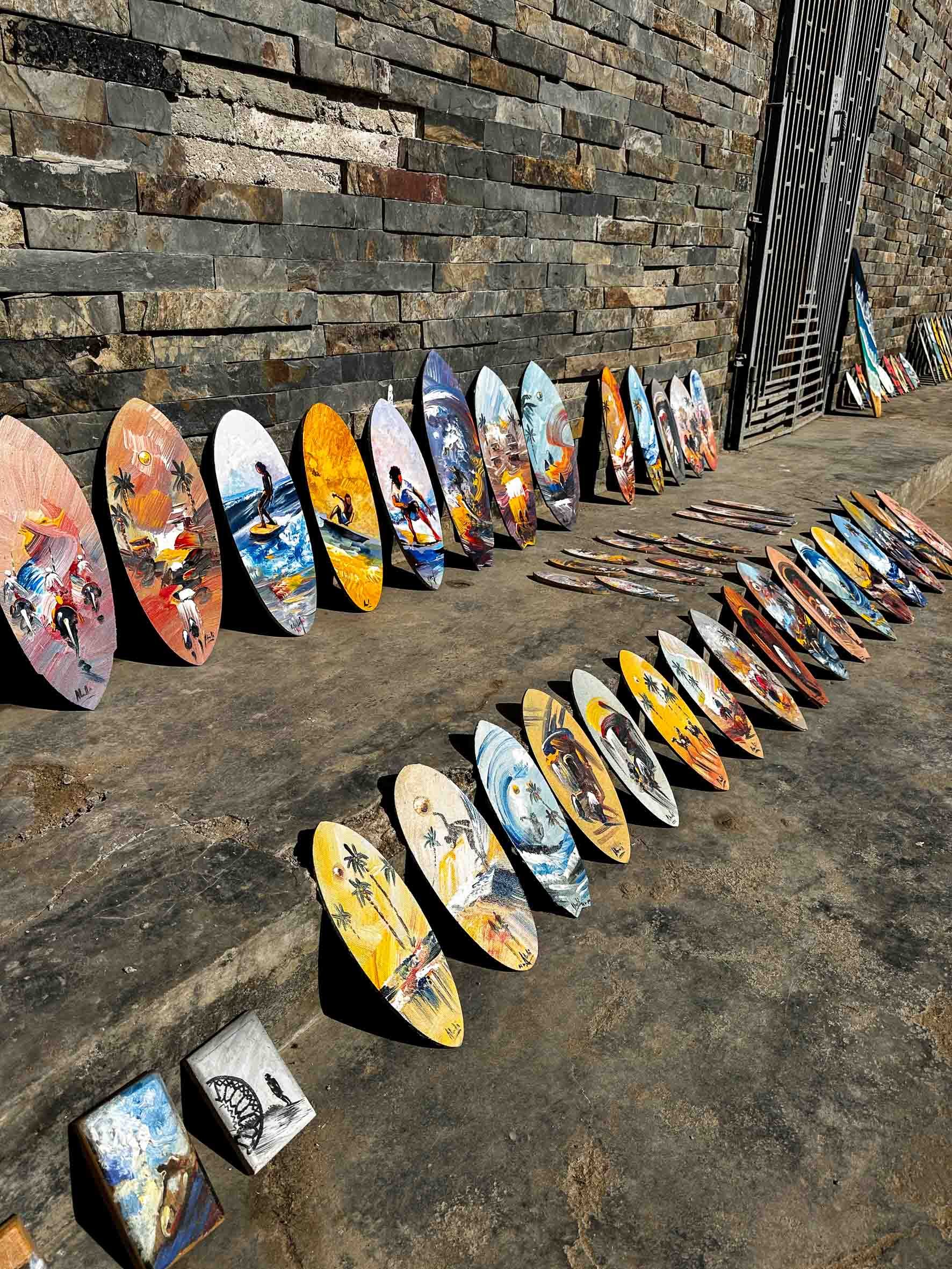 Display of colorful surfboards and small paintings outdoors against a brick wall and pavement.