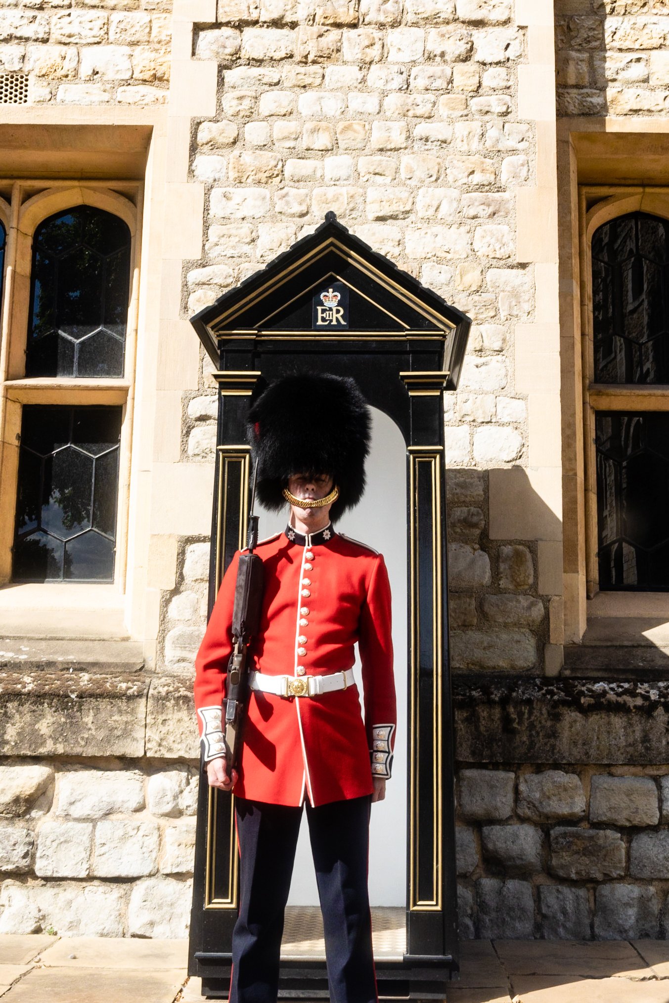 A British Royal Guard in uniform stands in front of a stone building with tall windows, wearing a red coat and a tall black bearskin hat.