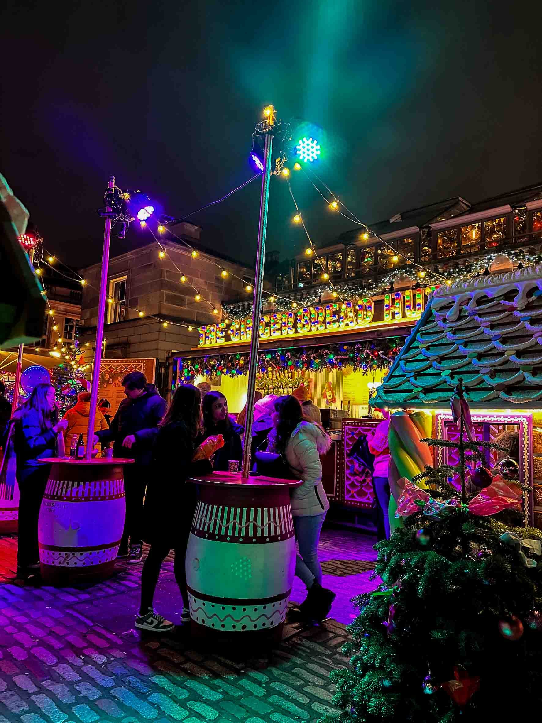 Festive outdoor scene at night with people gathered near a food stand decorated for Christmas, with a Christmas tree, colorful lights, and a sign reading 'The Gingerbread House'.
