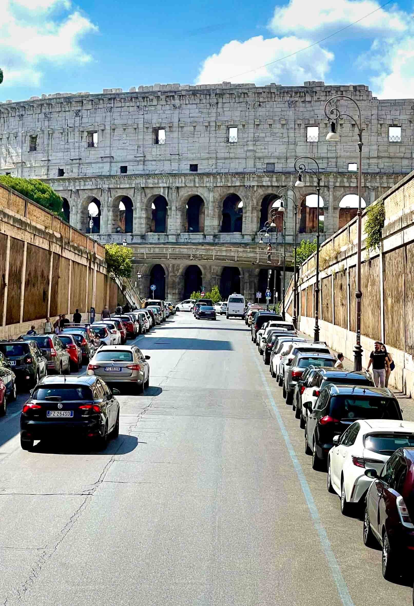 View of the Colosseum in Rome, Italy, with parked cars and pedestrians on the street in front of it under a partly cloudy sky.