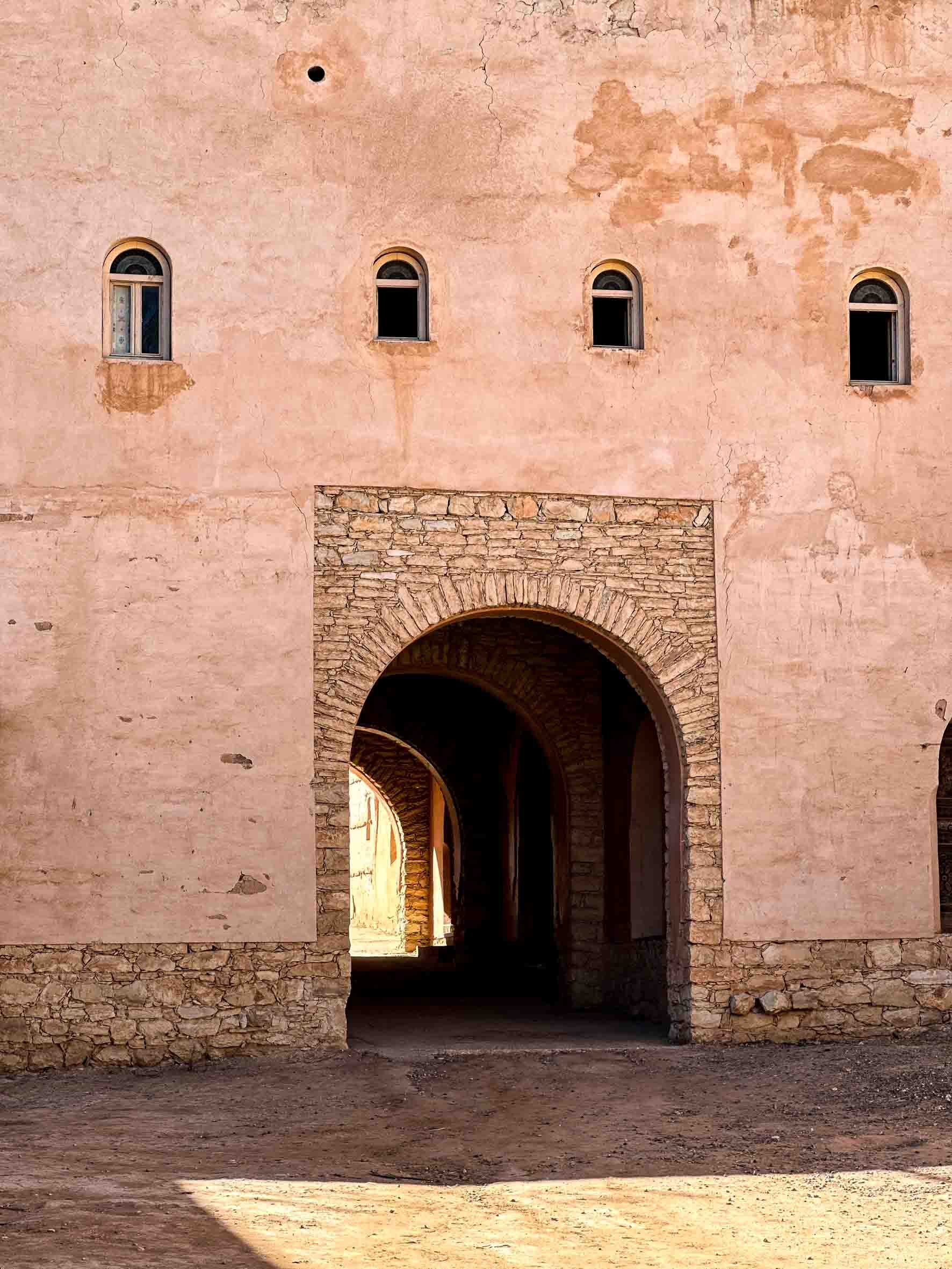 A pink stucco building with four small arched windows and a large arched entrance, with a dirt pathway in front.