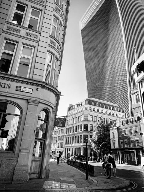 Black and white photo of a city street with historic and modern buildings, people walking, and trees lining the sidewalk.