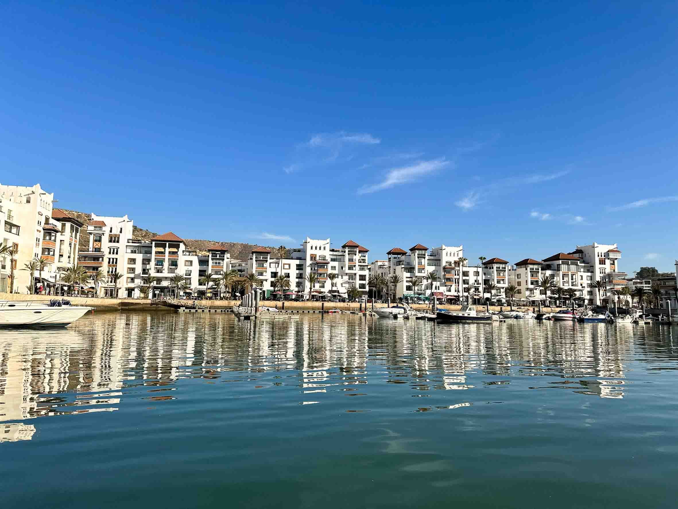 A marina with calm water reflecting white multi-story buildings and boats, under a bright blue sky with a few clouds.