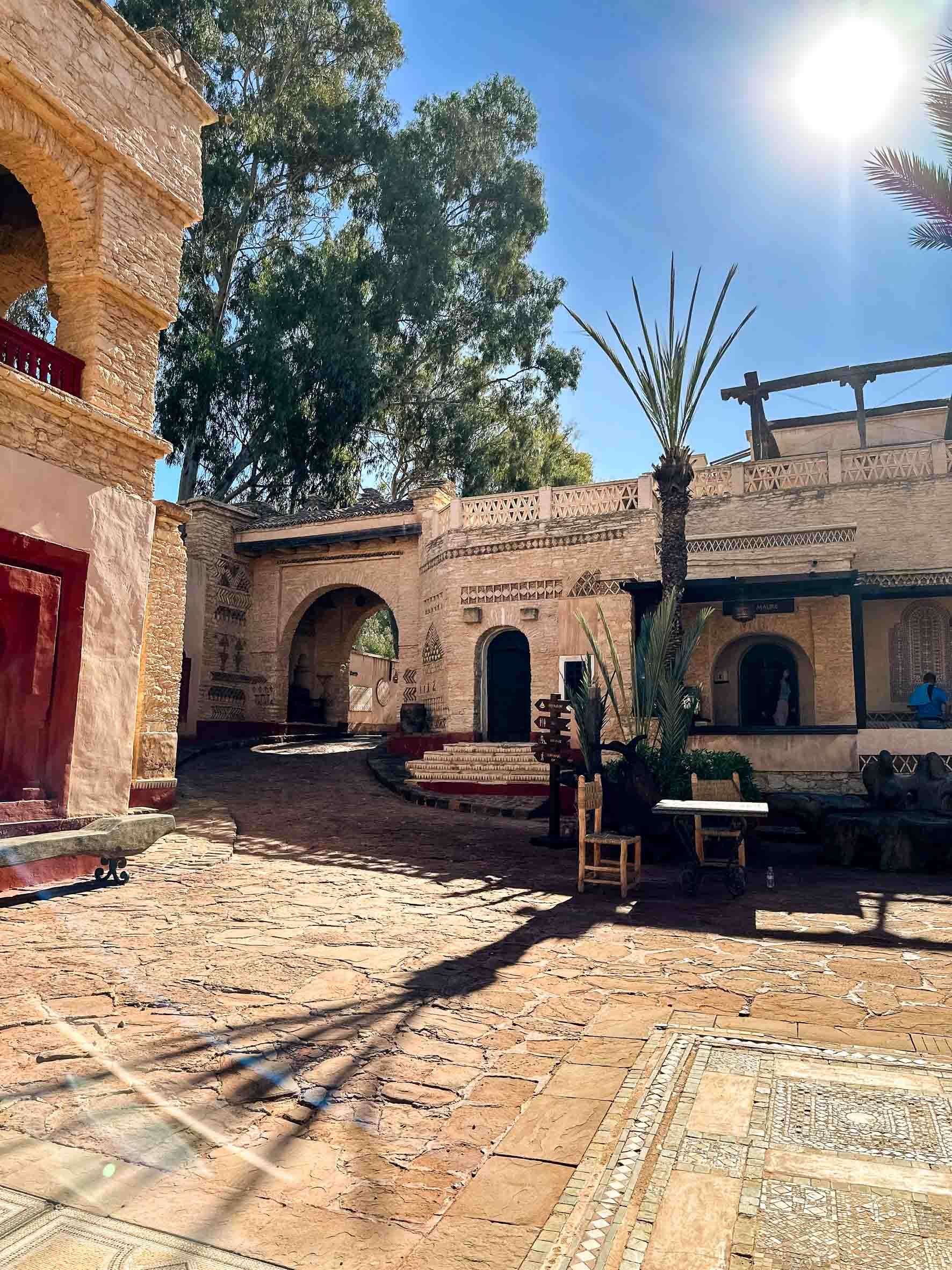 Sunlit courtyard with Spanish-style architecture, stone pavement, palm trees, and large trees in the background.