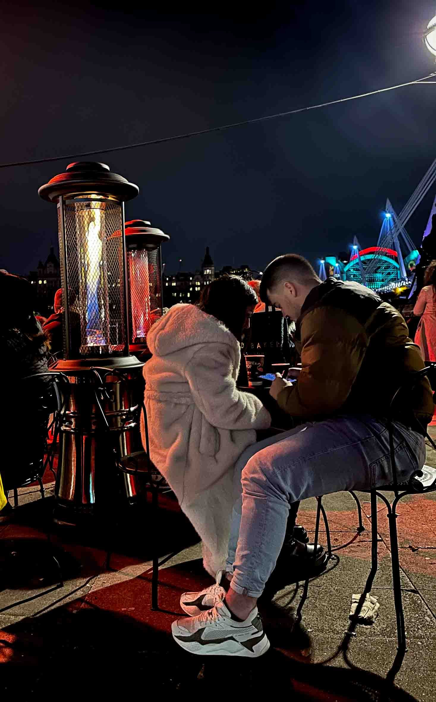 Two people sitting at an outdoor table at night, illuminated by a tall patio heater, with a city skyline and amusement park rides in the background