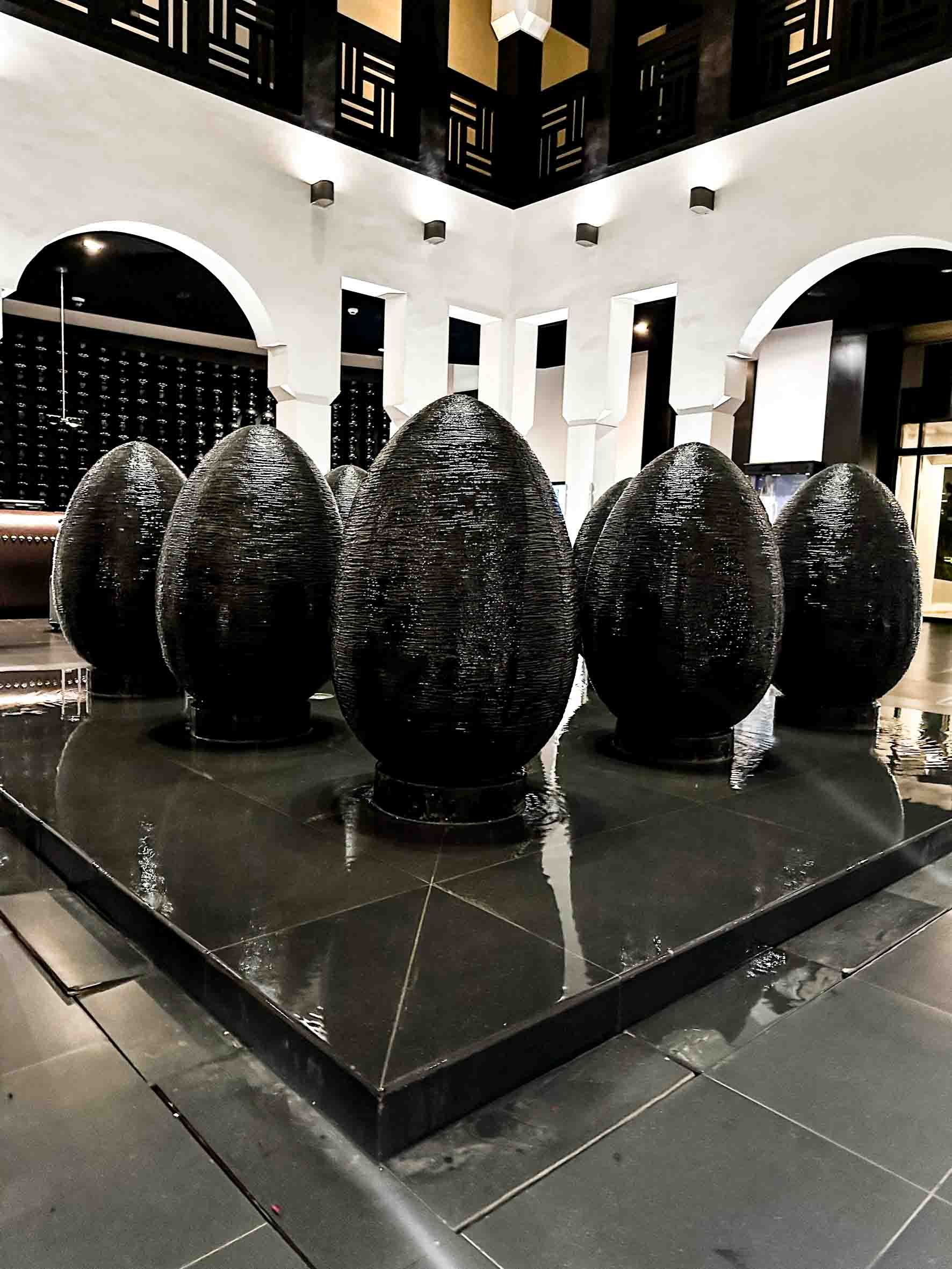 Six large black eggs arranged on a black platform inside a modern hotel lobby with white walls, arches, and decorative lighting.
