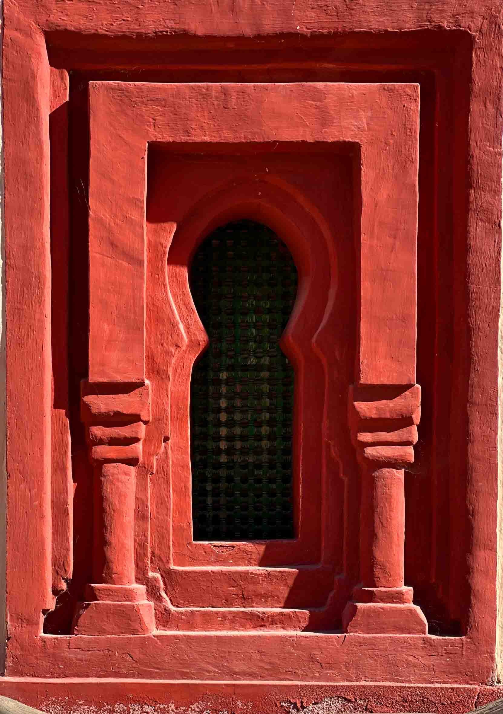 A red stone window frame with a keyhole-shaped opening, featuring decorative columns on each side and a dark, patterned interior visible through the opening.