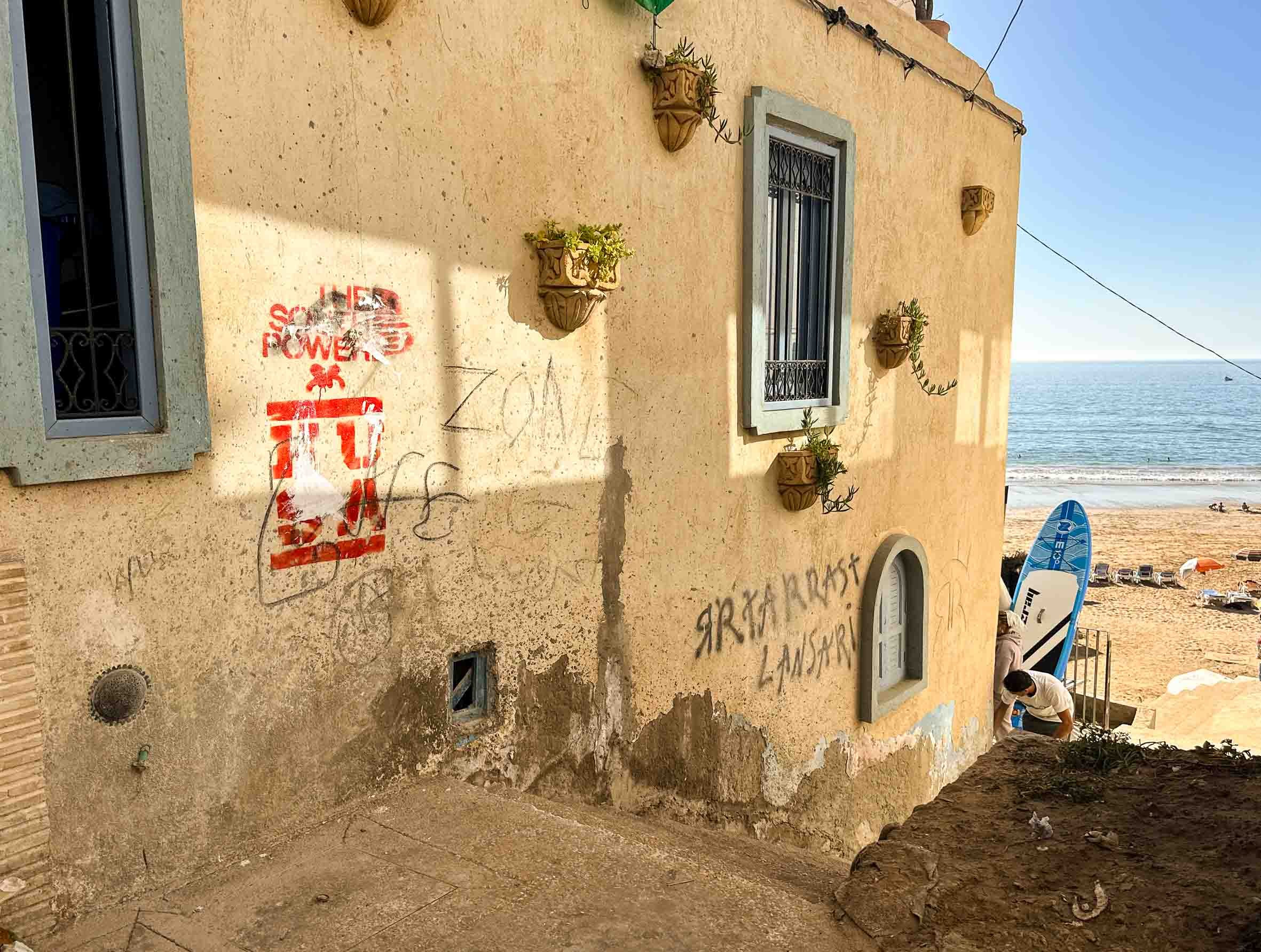A building with a yellow textured wall decorated with potted plants, two windows with black metal bars, and graffiti. A person bends down near the bottom right, with a beach and ocean visible in the background.