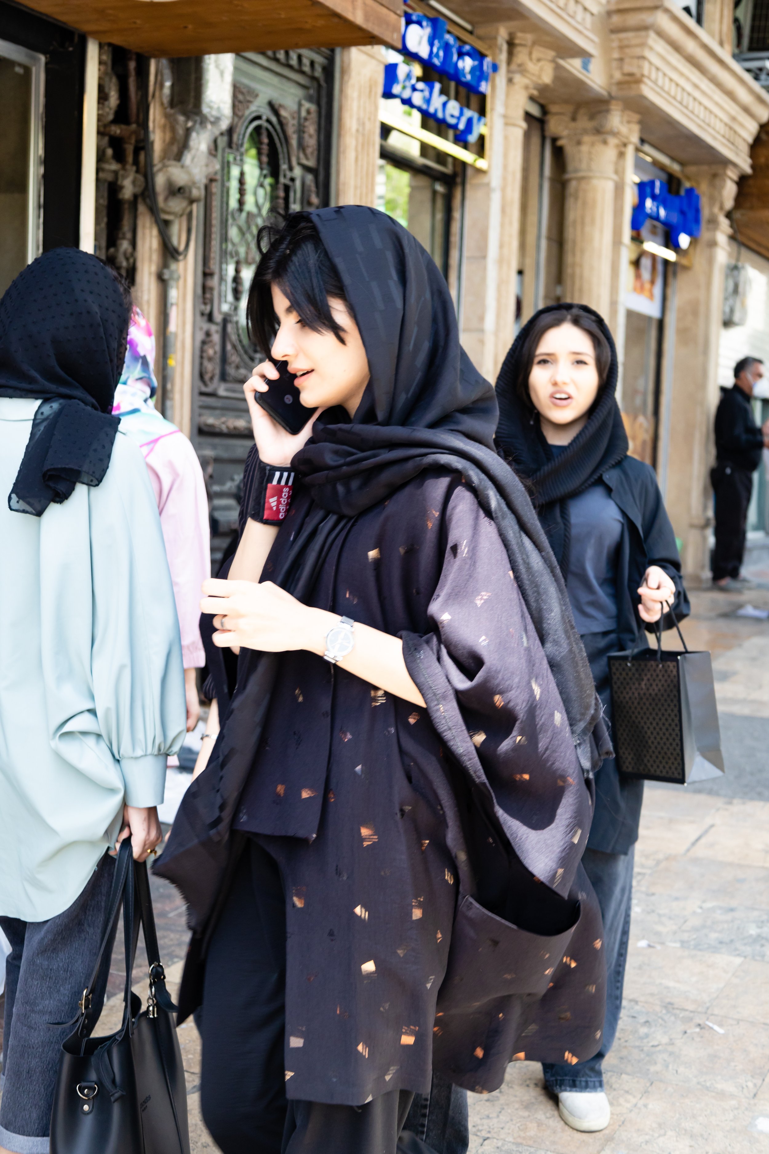 Group of women walking on a city sidewalk, with one woman talking on her mobile phone and wearing a black headscarf.